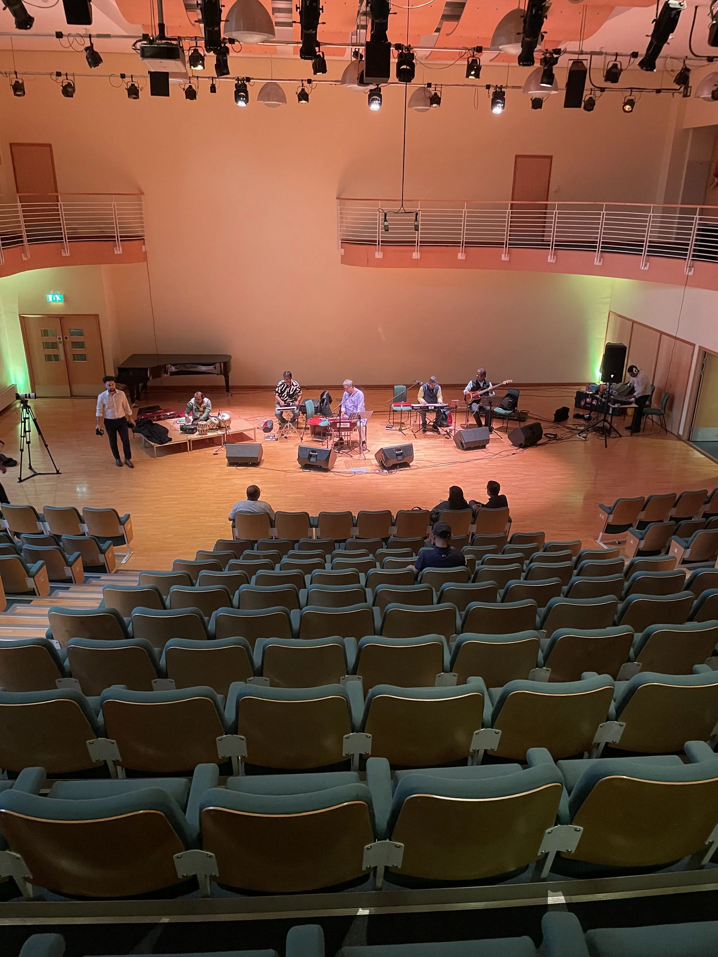 A concert hall stage with seven musicians preparing for a performance. The stage is equipped with various instruments, microphone stands, and sound equipment. The hall has an audience sitting in rows of chairs facing the stage.