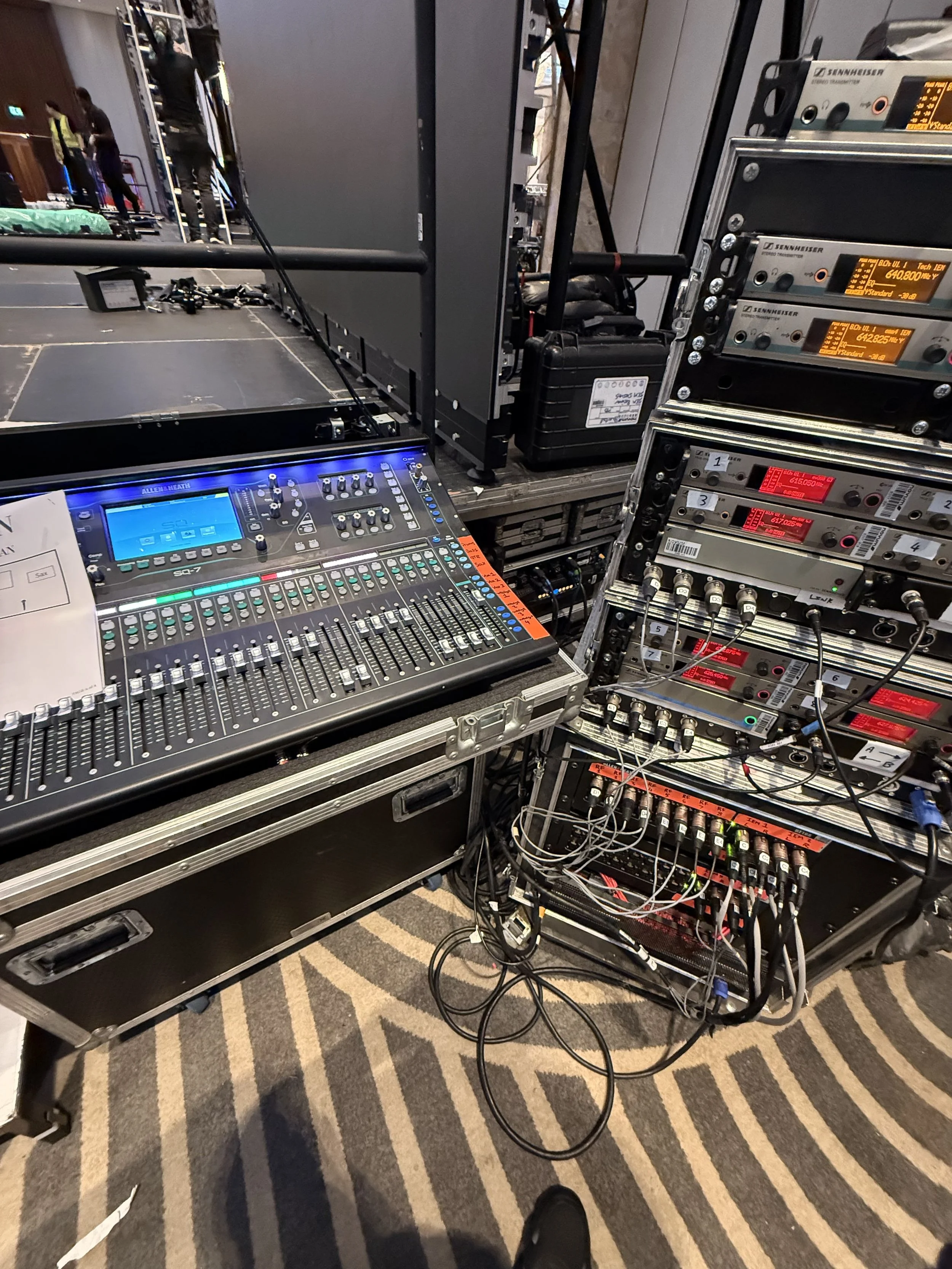 A stage setup with audio equipment, including a mixing console, power amplifiers, and various cables. There are people in the background preparing the stage, and the scene is indoors.