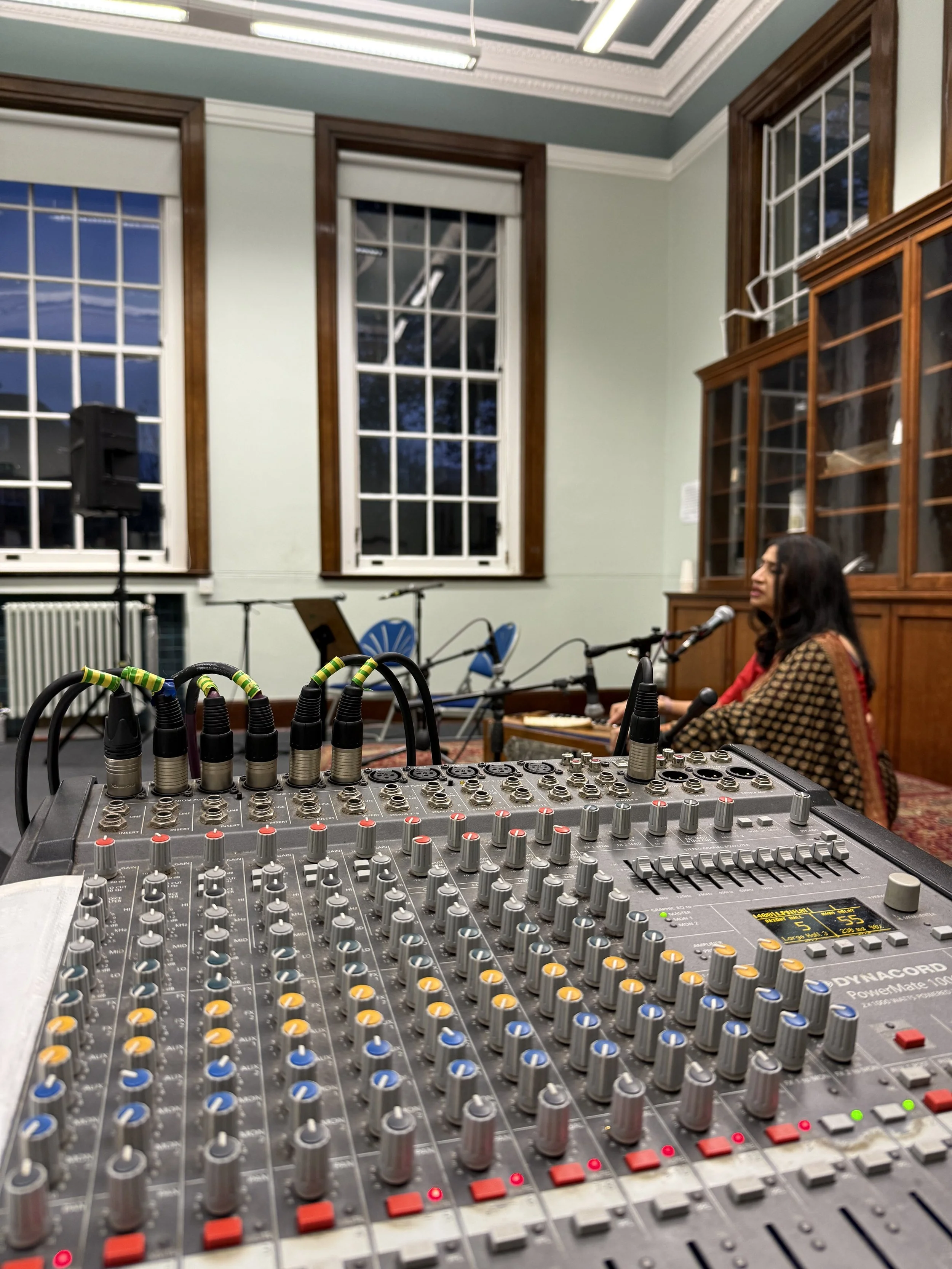 Close-up of a professional audio mixing console with various knobs, sliders, and colored buttons, set up in a room with large windows and wooden accents.