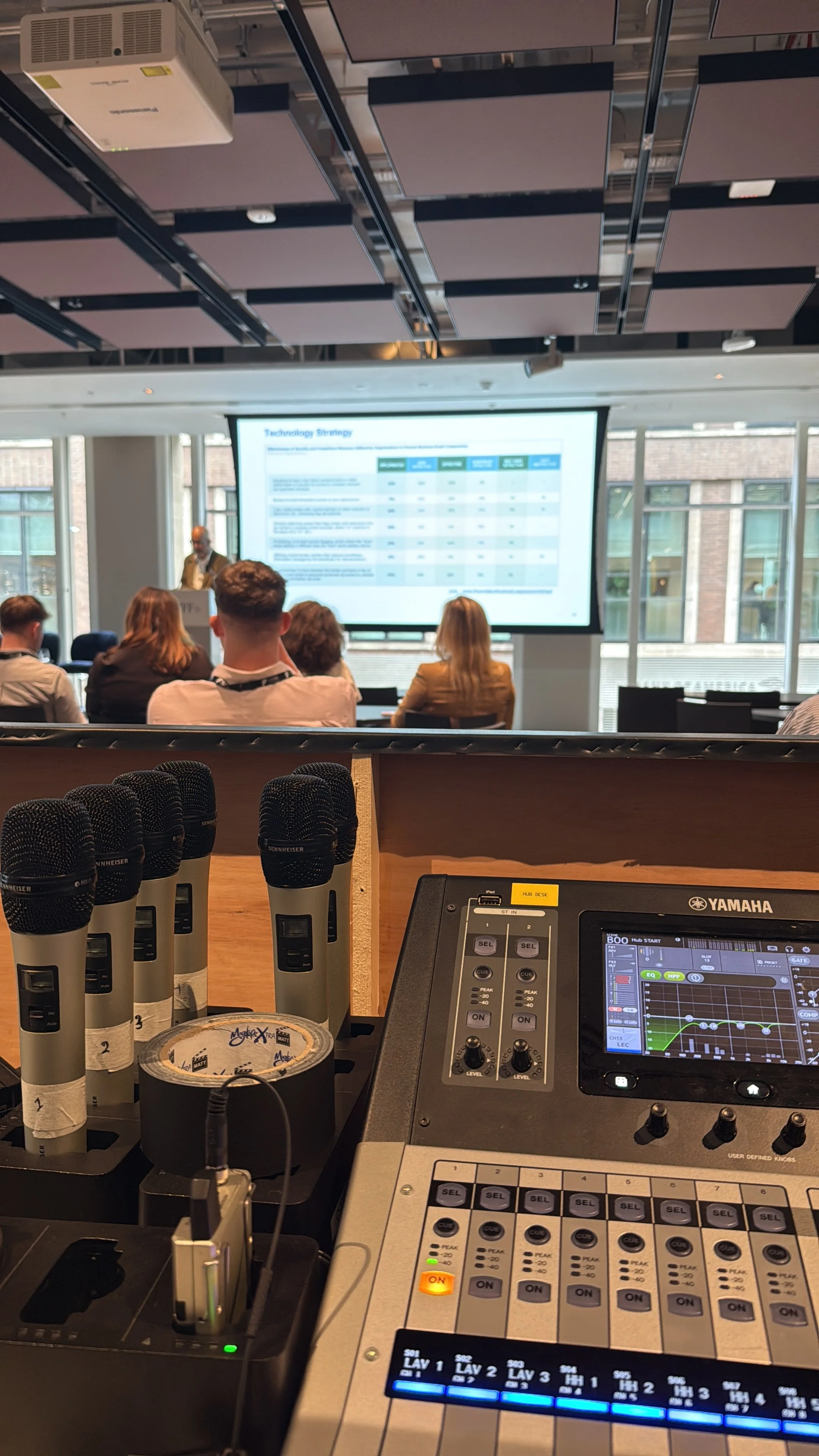 View from a DJ booth showing microphones, audio equipment, a stage monitor, and a presentation screen with an audience watching a speaker in a conference room.