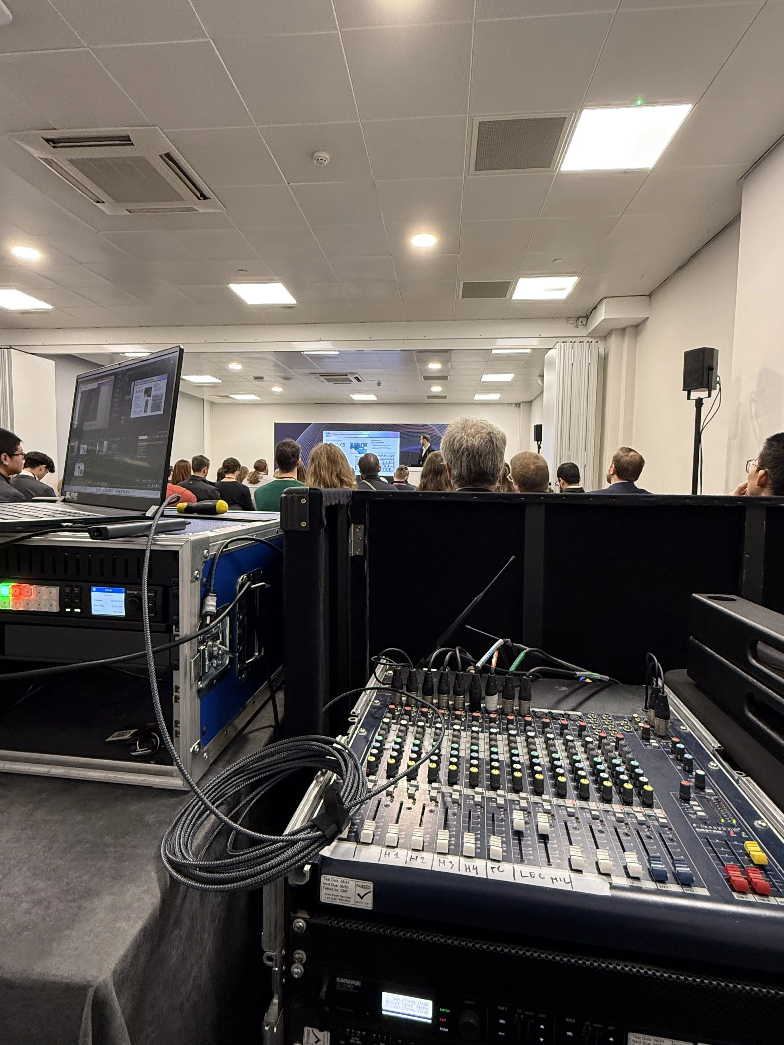 Conference room with a presentation screen at the front, attendees sitting at tables, and audio/visual equipment in the foreground.
