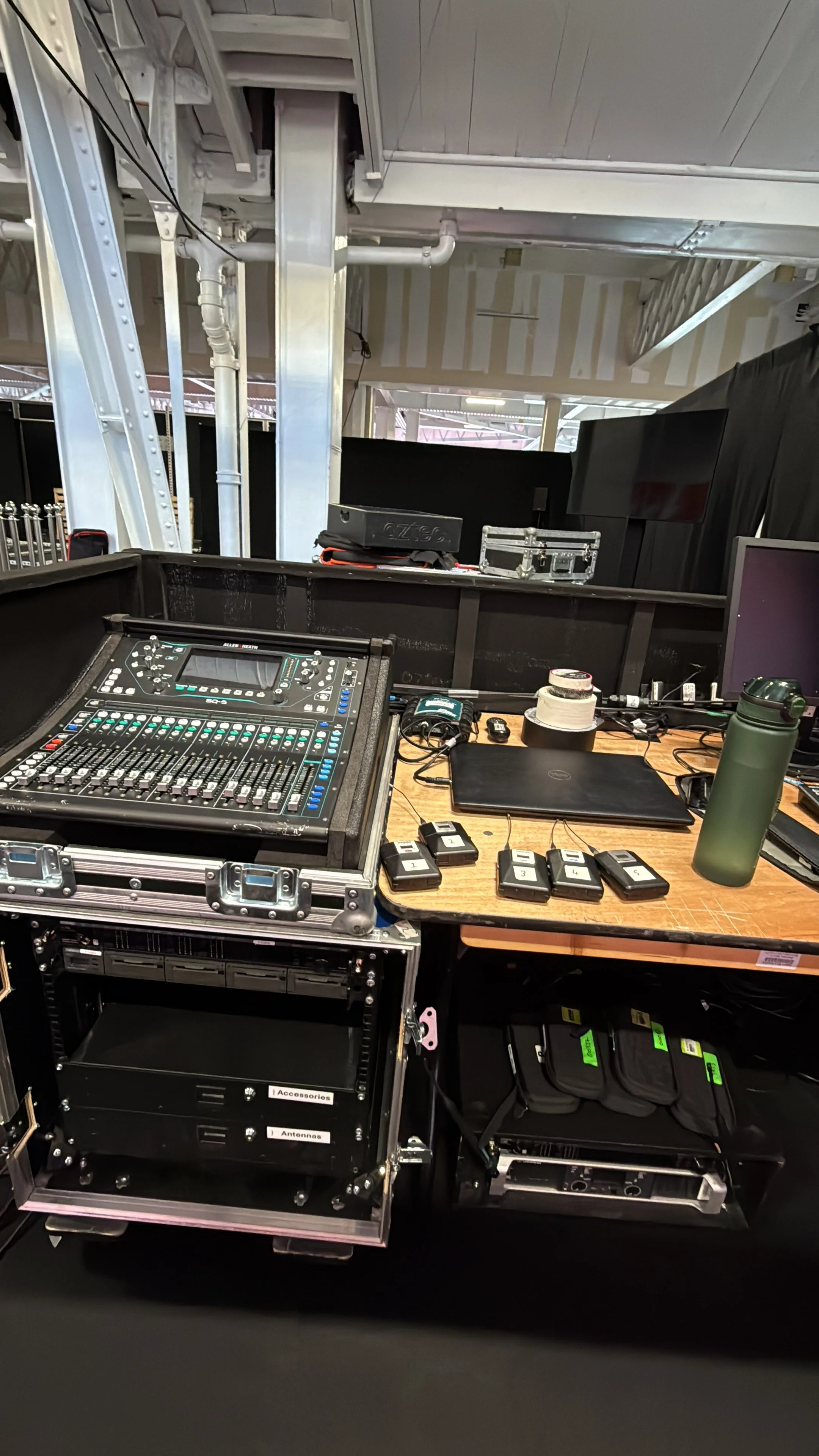 A control booth with audio mixing equipment, laptops, key fobs, a water bottle, and other electronic devices. In the background, there are structural elements and black curtains.