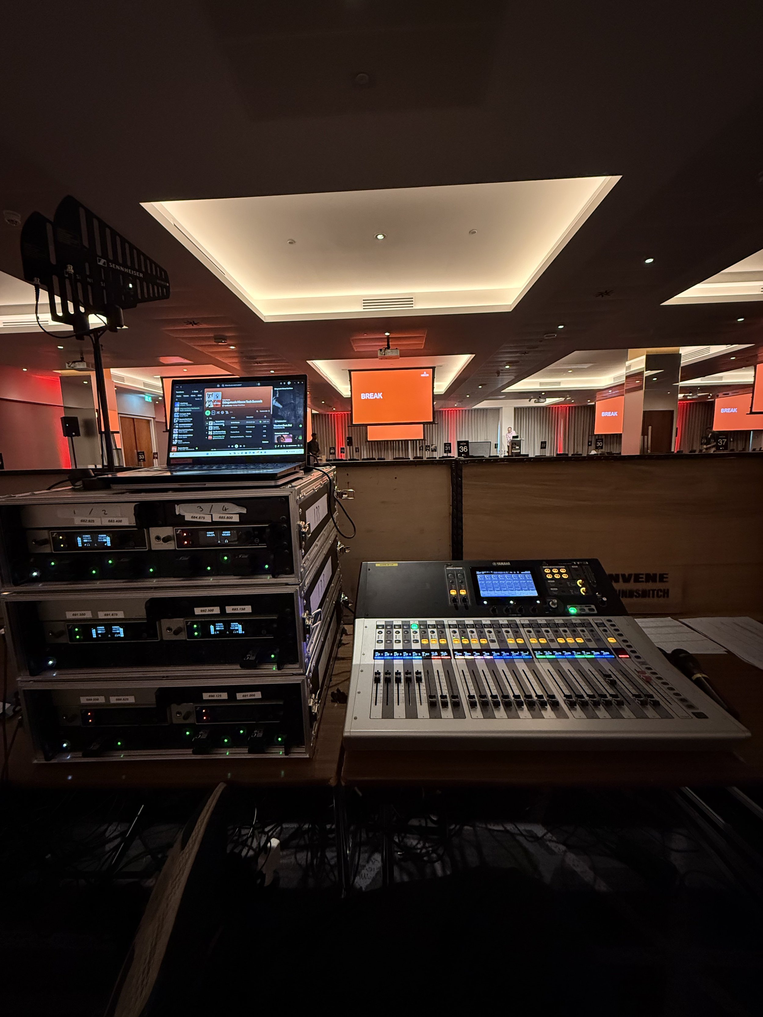 View of a conference or event hall with a stage and large screens displaying the word 'BREAK'. In the foreground, audio and lighting equipment, including a laptop, audio mixer, and other electronic devices, suggests preparation for a presentation or 