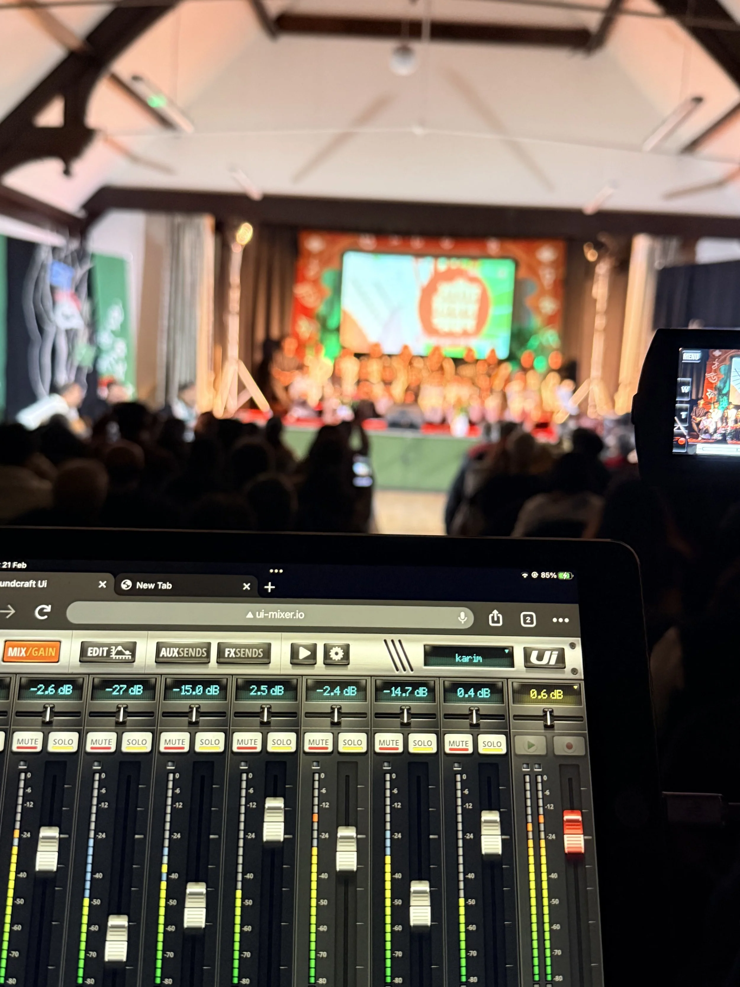 Close-up of a digital audio mixing console with sliders and meters, in a dimly lit room with a blurred stage and audience in the background.