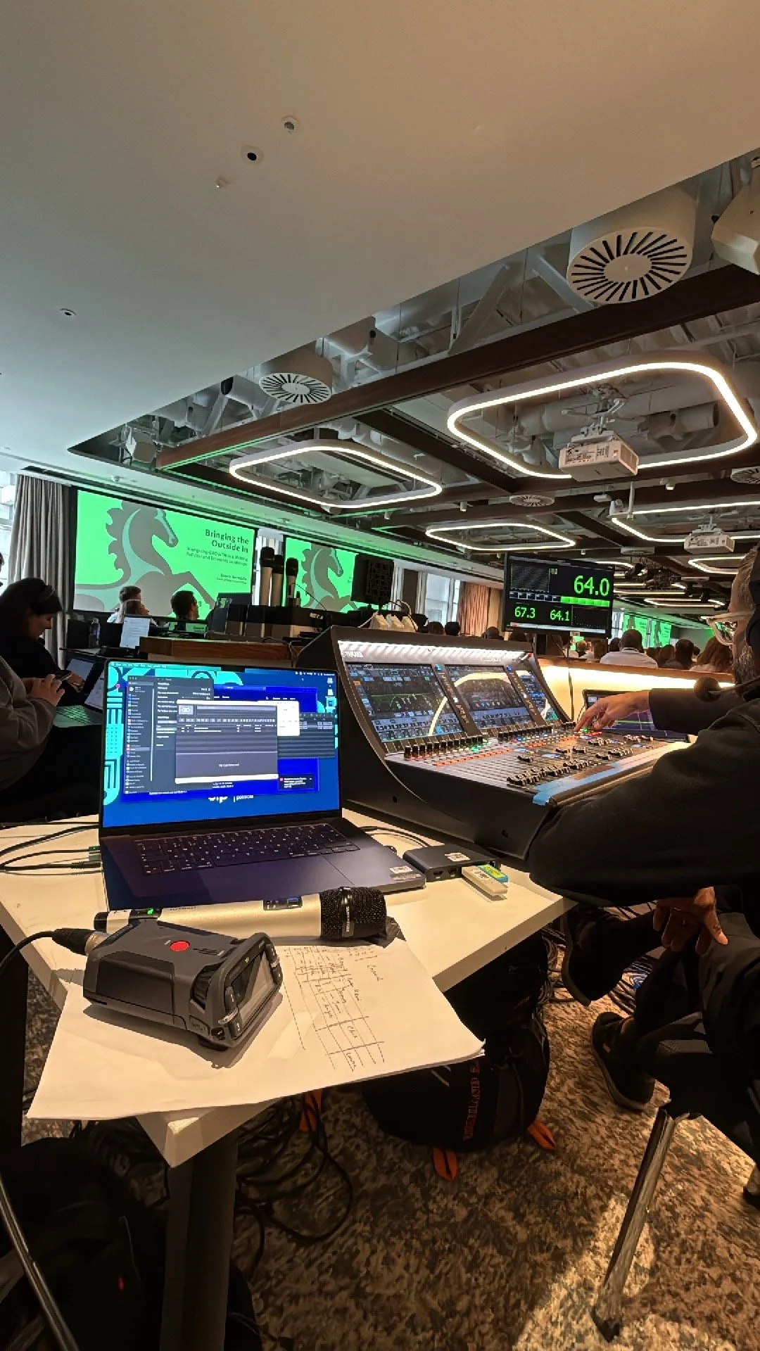 Conference room with audio and visual equipment, including laptops, audio mixer, microphones, and large digital screens displaying graphs and information. Several people are working or observing, with a presentation projected on the screen.
