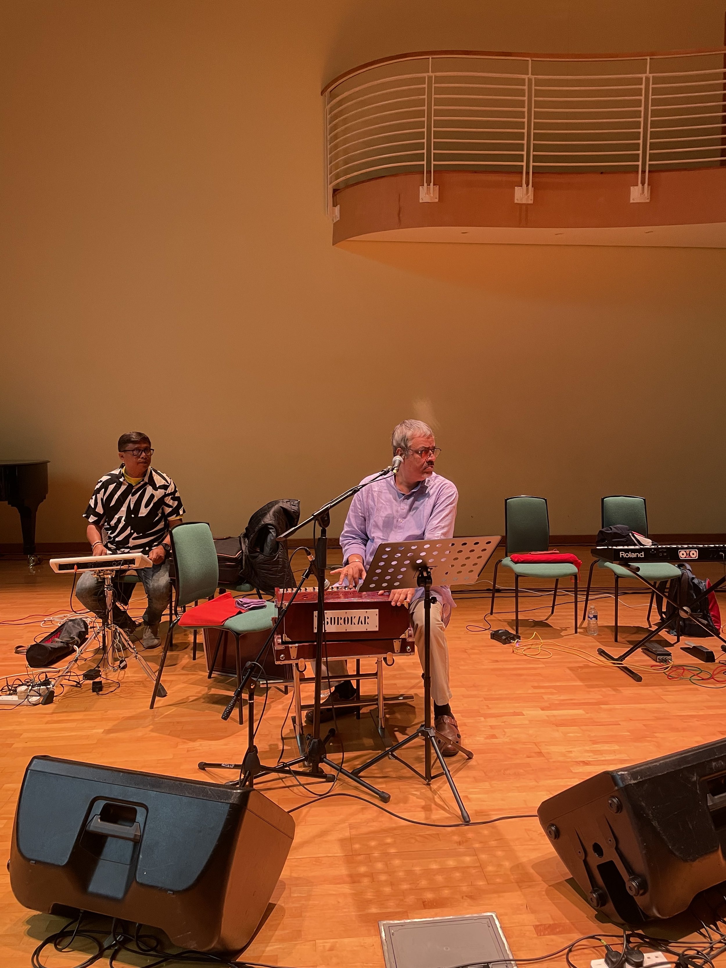 Two musicians performing on stage in a music venue. One is playing a keyboard, and the other is sitting in front of a music stand with a percussion instrument. There are large speakers and various audio equipment on the stage.