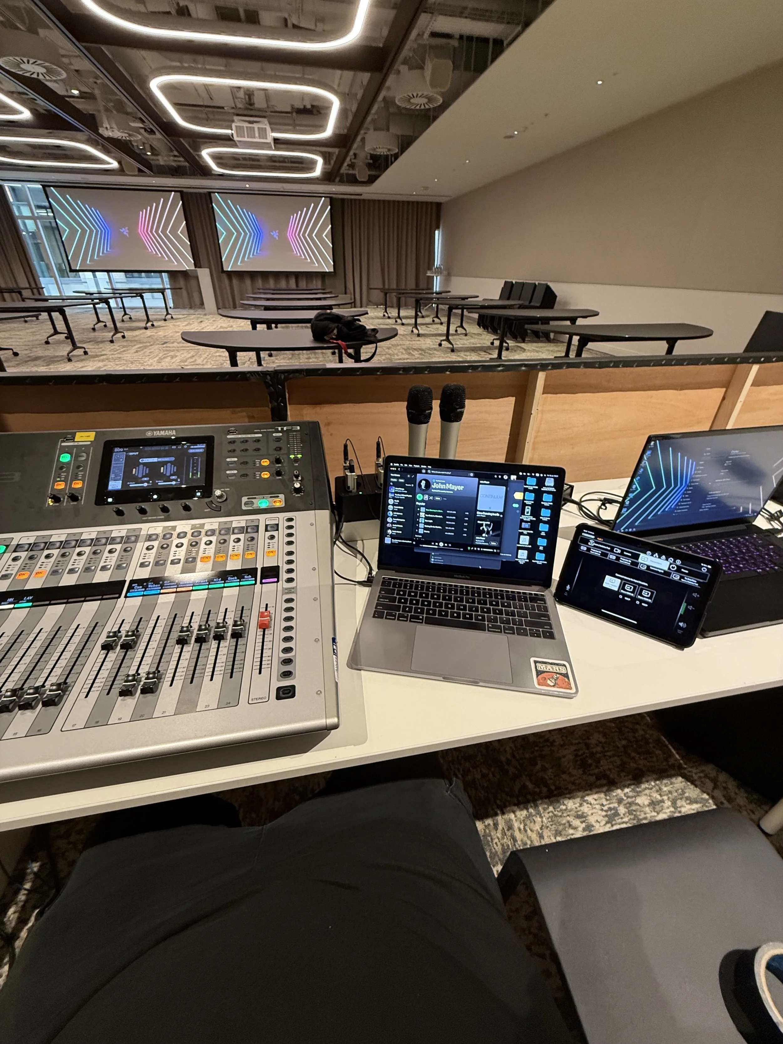 A sound and lighting control booth with a mixing console, laptops, microphones, and a person sitting in front of the equipment. The auditorium has rows of chairs and two large screens displaying colorful geometric digital designs.