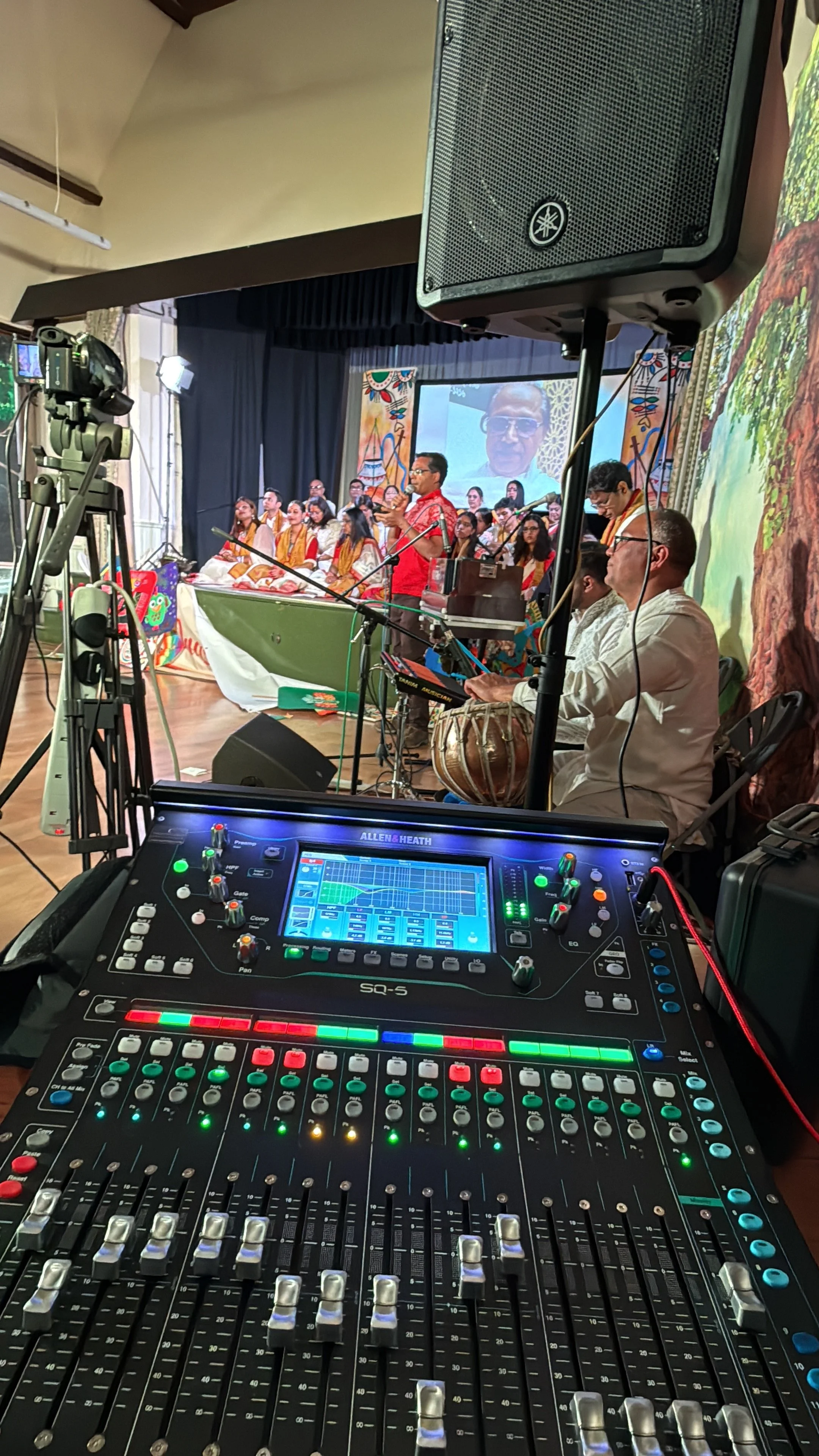 View from behind a sound mixer at a stage performance. The mixer's controls are visible in the foreground, with people on stage in the background, including a man in a red shirt speaking into a microphone and a group of performers dressed in traditio