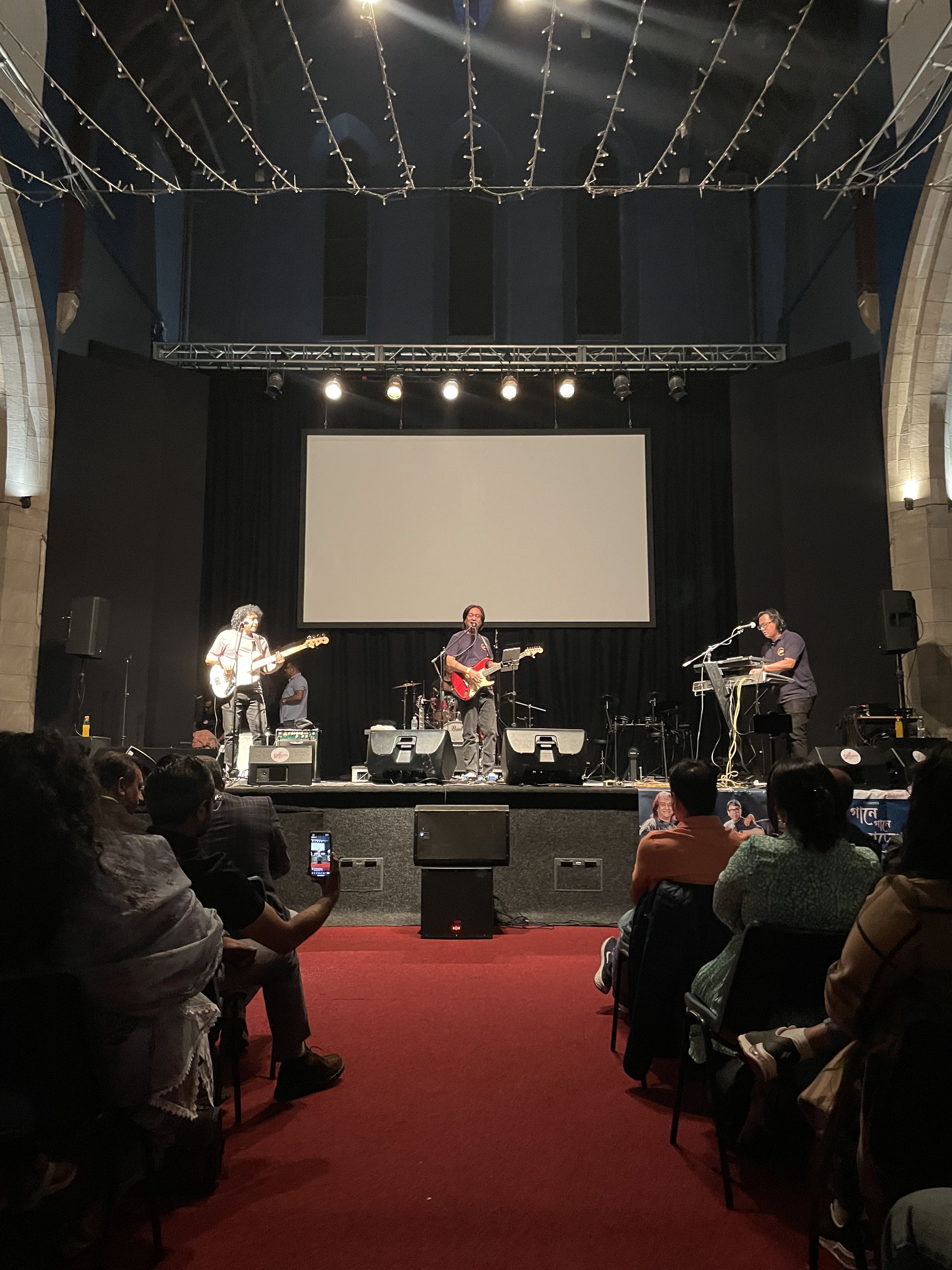 A band performing on a stage in front of an audience seated in chairs. The band includes a guitarist, keyboardist, and drummer, with a large white screen behind them. Audience members are taking photos or watching the performance.