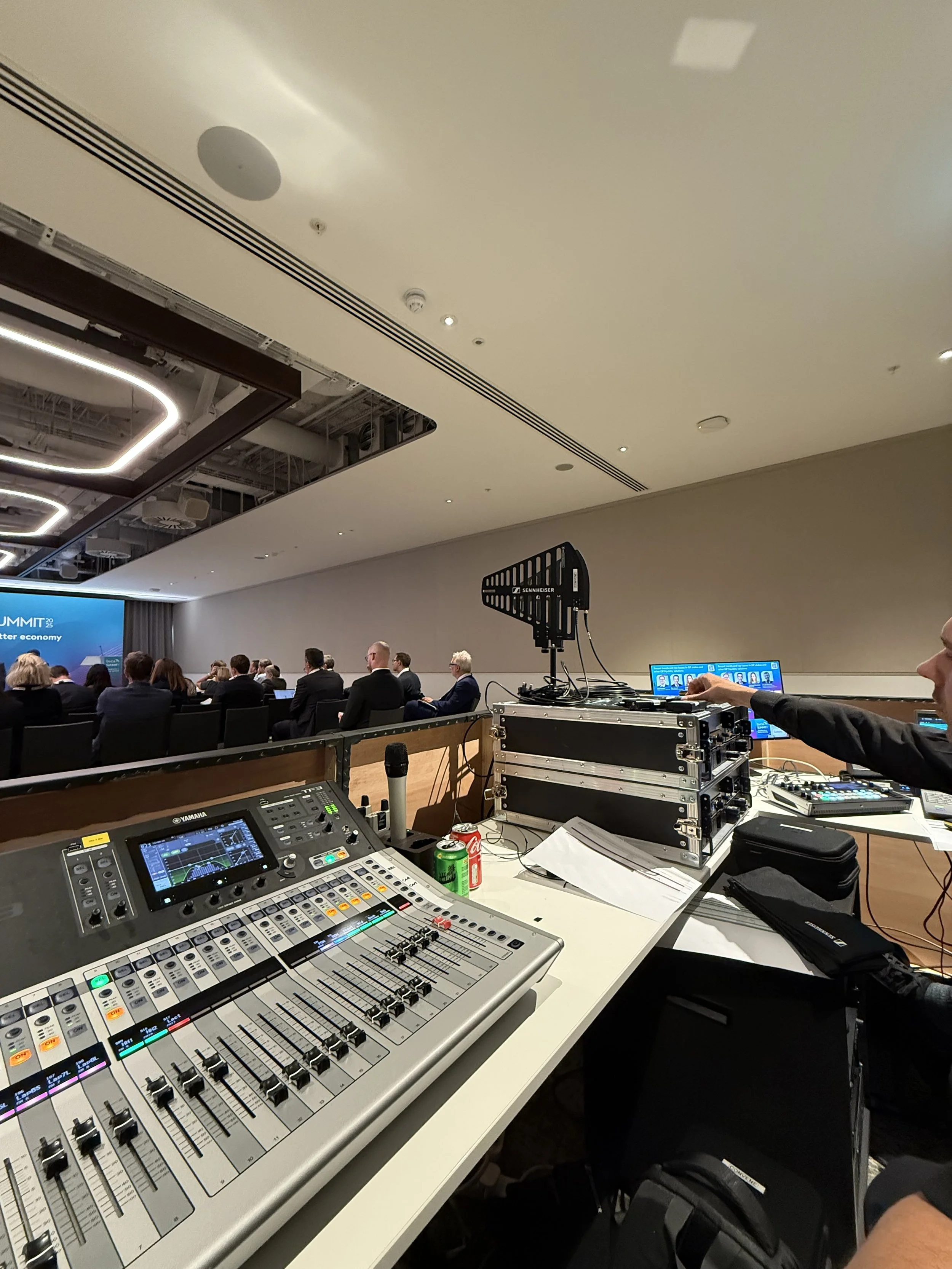 A conference room filled with people seated at tables, with audio and visual equipment at the forefront, including a large mixing console, a microphone, and various digital devices. The ceiling has modern lighting fixtures and vents, and a large scre
