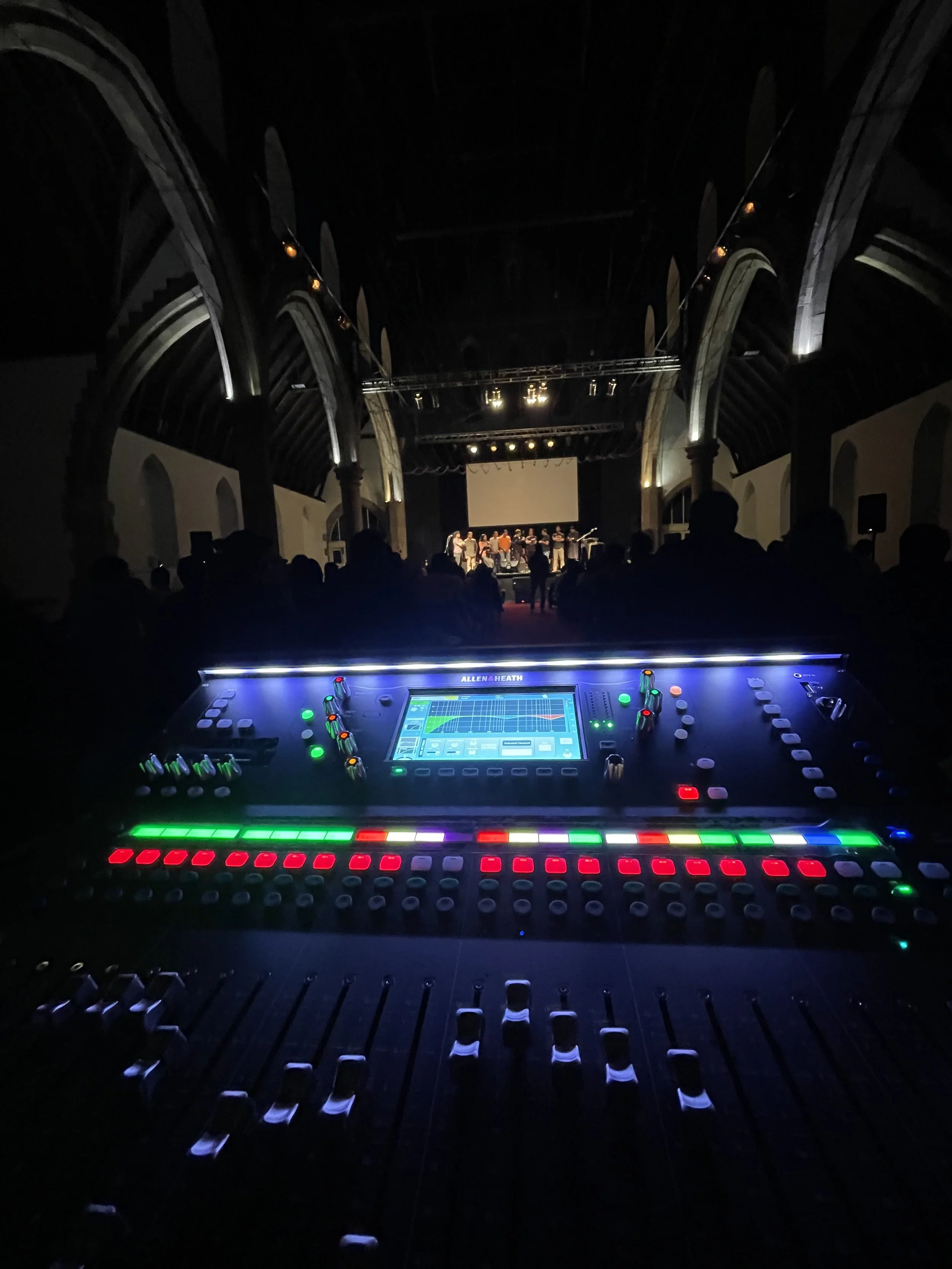 A stage with performers and a large screen in a dark concert hall, with an audio mixing console in the foreground.