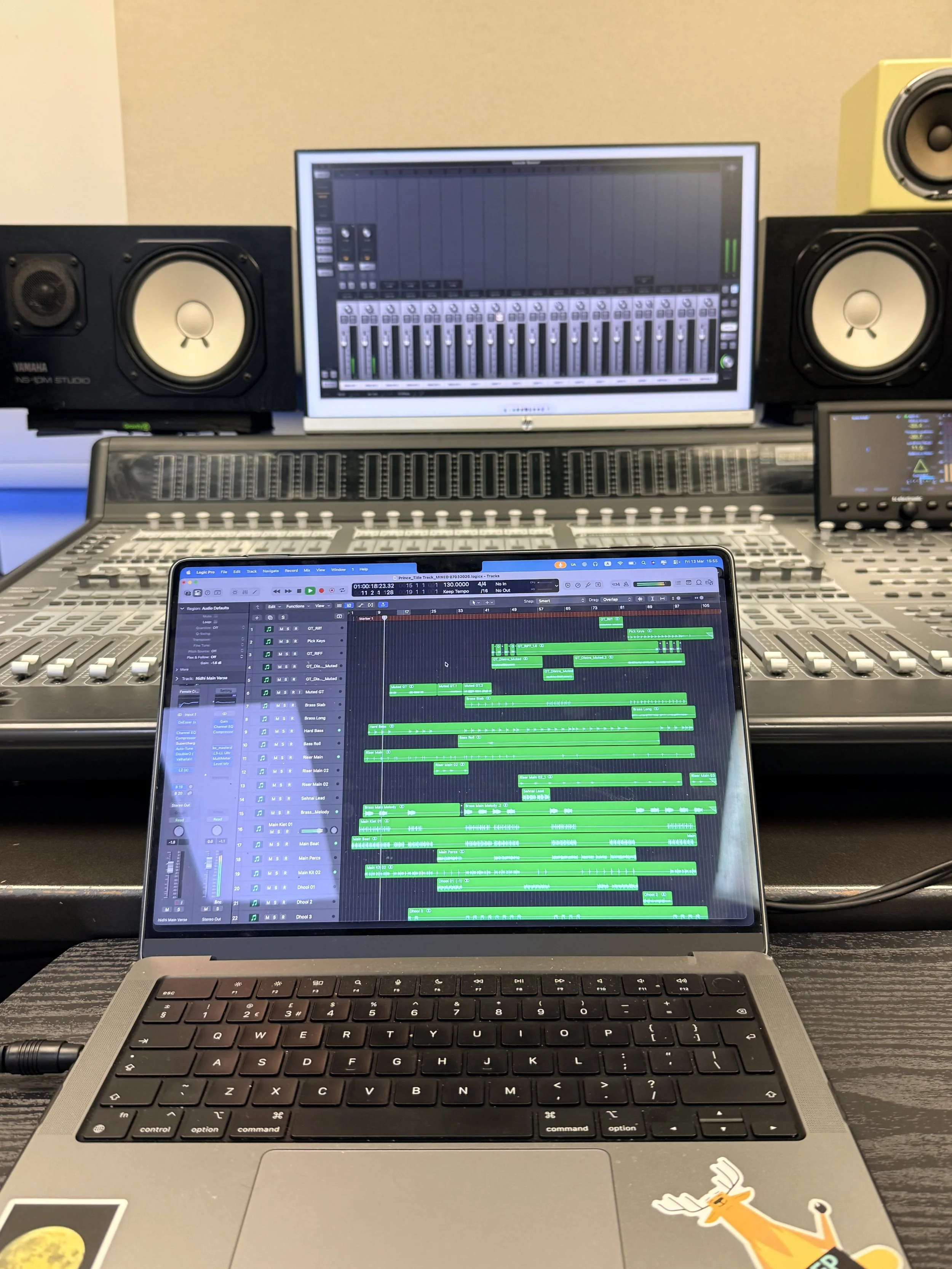 A music production studio setup with a laptop, mixing console, two studio monitor speakers, a computer screen with digital audio workstation, and an additional small screen, with a decorative sticker of a cartoon deer on the laptop.