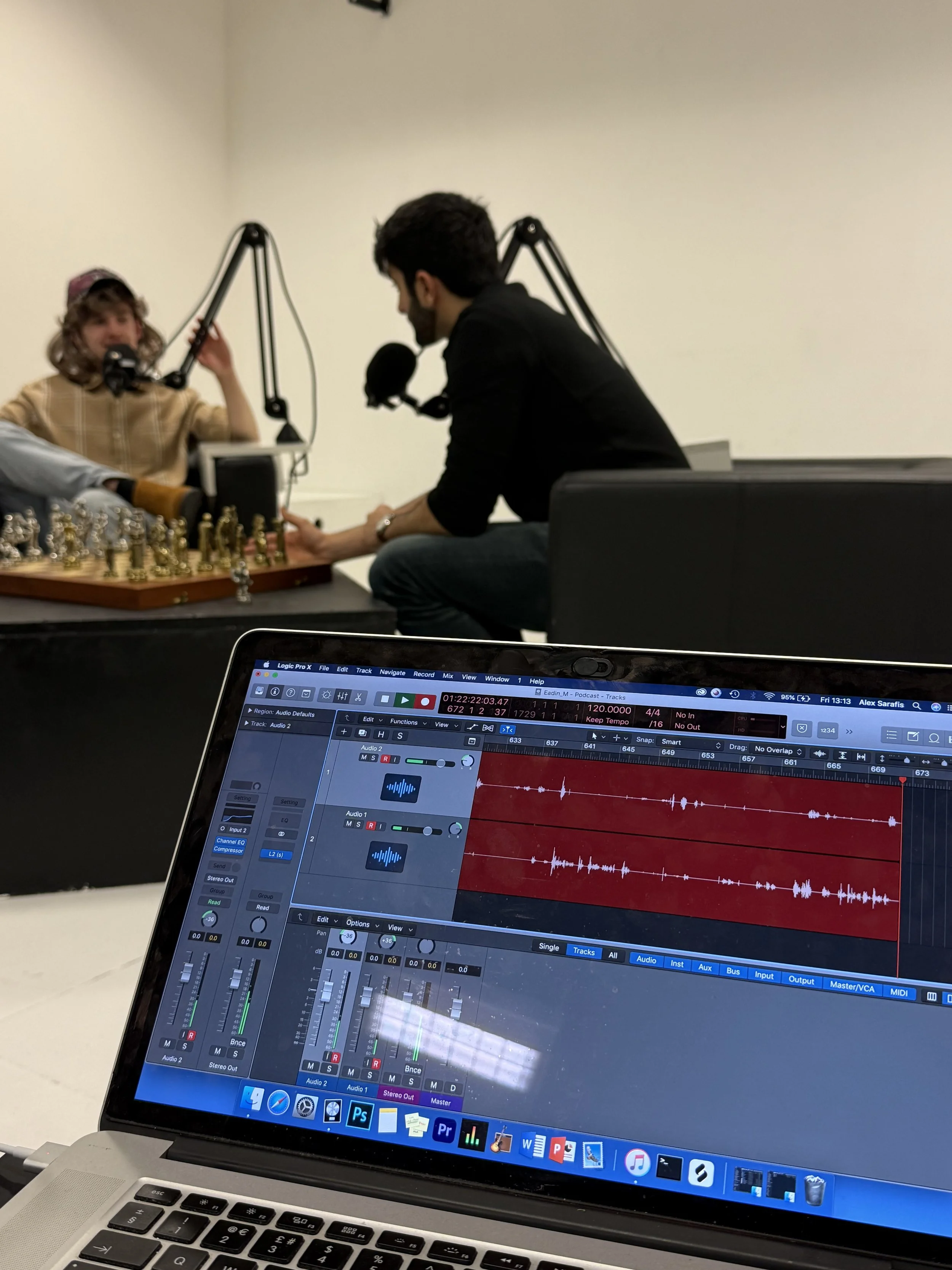 A laptop screen showing digital audio workstation software with multiple tracks and waveforms. In the background, a person is sitting at a table with a microphone on a boom arm, engaged in a podcast or audio recording, with another person in front of