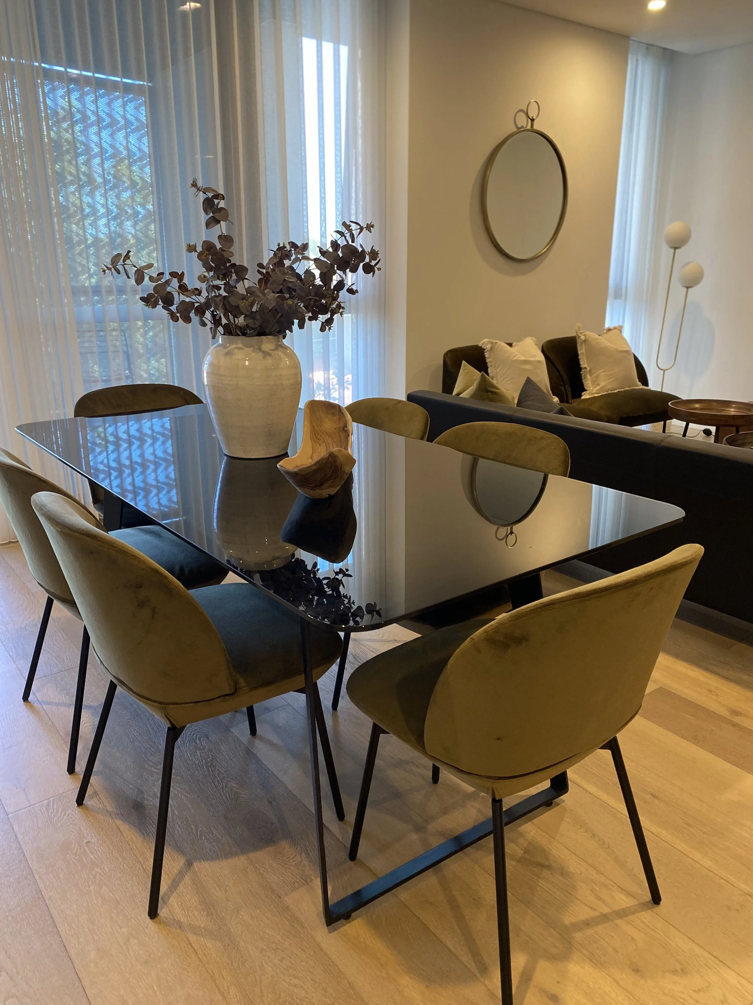 A dining area with a black glass table, six yellow chairs, a large vase with dried eucalyptus on the table, and a living room with black and beige sofas, pillows, a round mirror, and floor lamps, near large windows with sheer curtains.