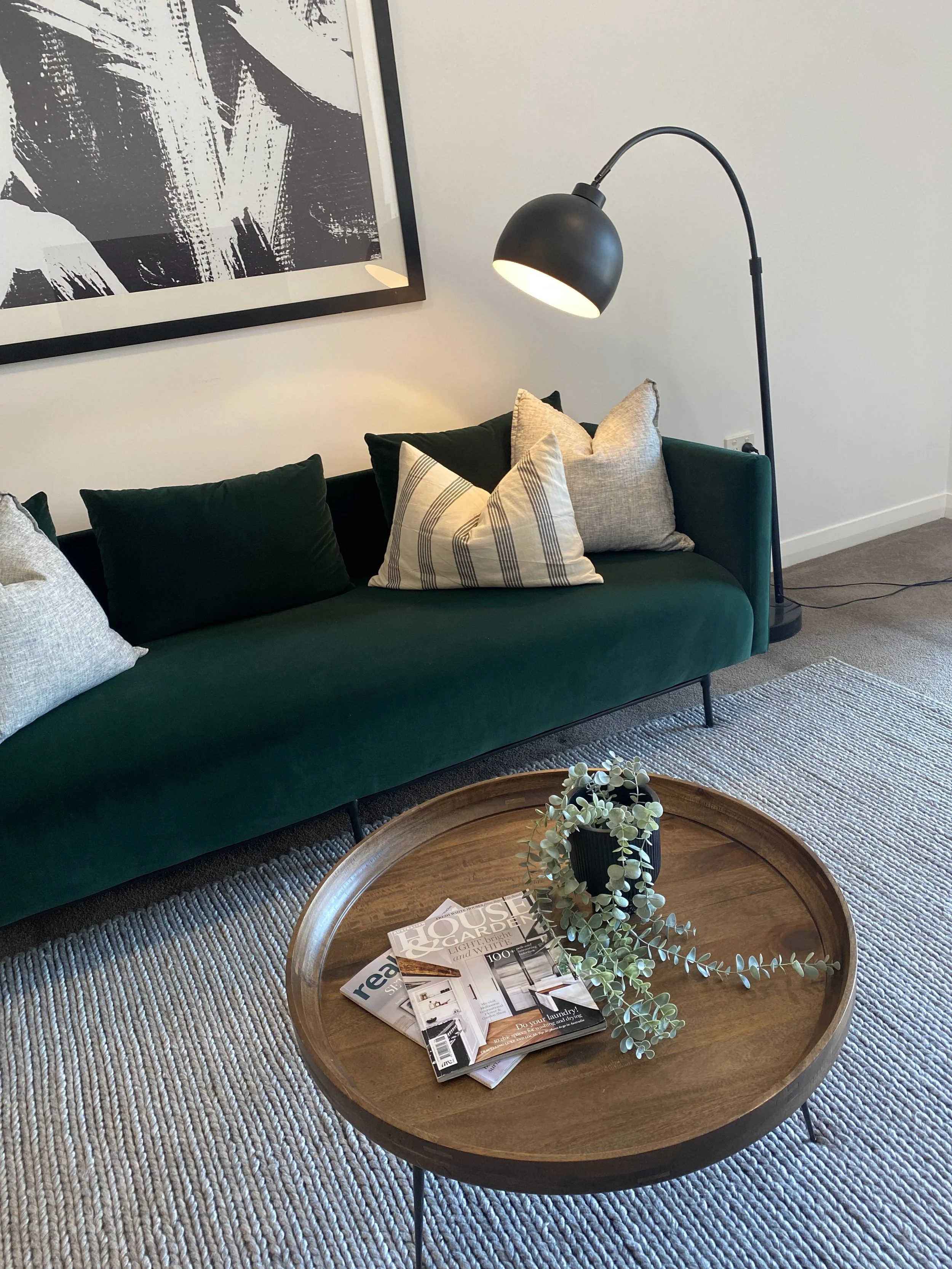 A living room corner with a green velvet sofa, patterned and solid pillows, a black arc floor lamp, a round wooden coffee table with a plant and magazines, a textured rug, and a large black and white abstract art piece on the wall.