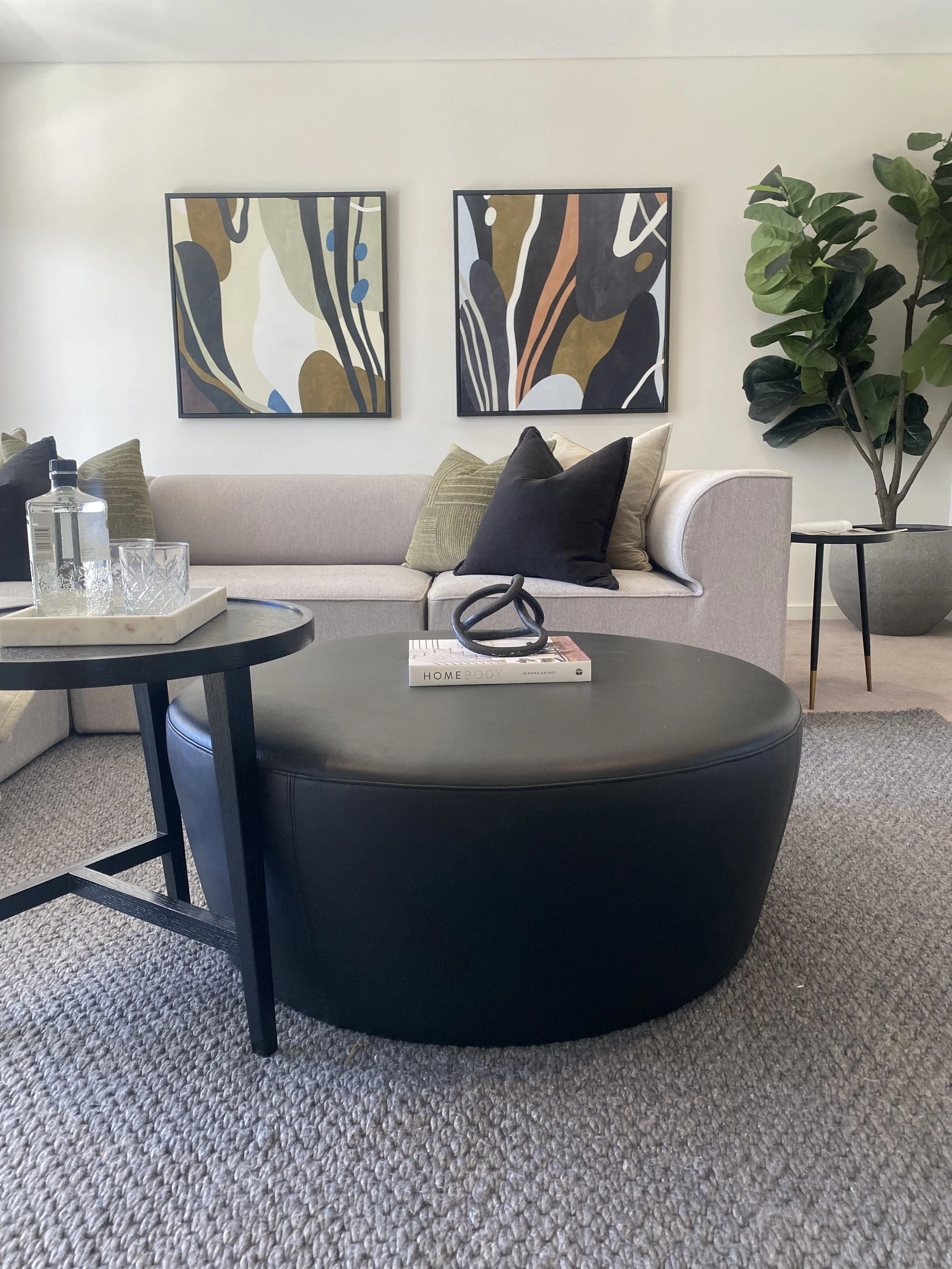 Modern living room with beige sofa, black and green throw pillows, black oval ottoman with decorative object and book, black side tables with glass tray and bottles, abstract wall art, large potted plant, neutral-toned carpet.