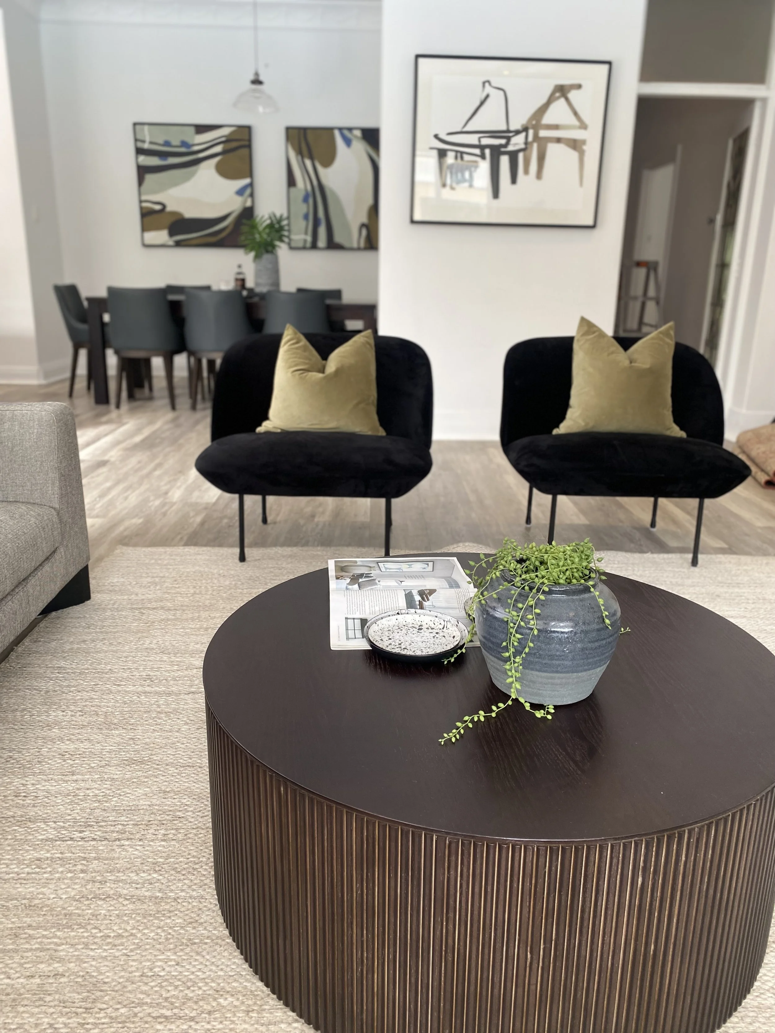 Living room with black armchairs with beige pillows, a round wooden coffee table with a potted plant and magazine, and abstract artwork on the walls.