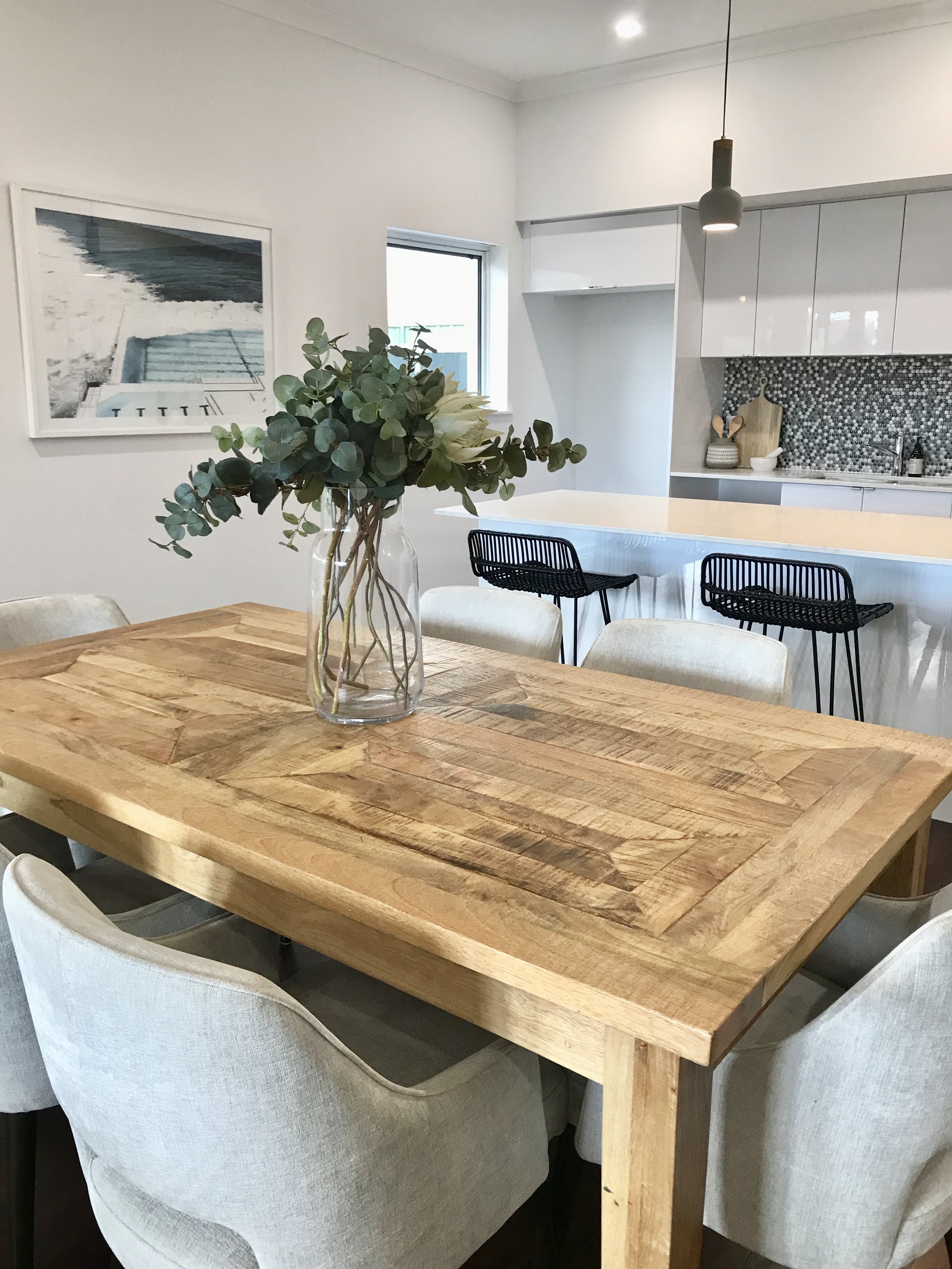 A dining area with a wooden table decorated with a glass vase with eucalyptus branches. There are beige upholstered chairs around the table, and a kitchen with a white island and black barstools in the background.