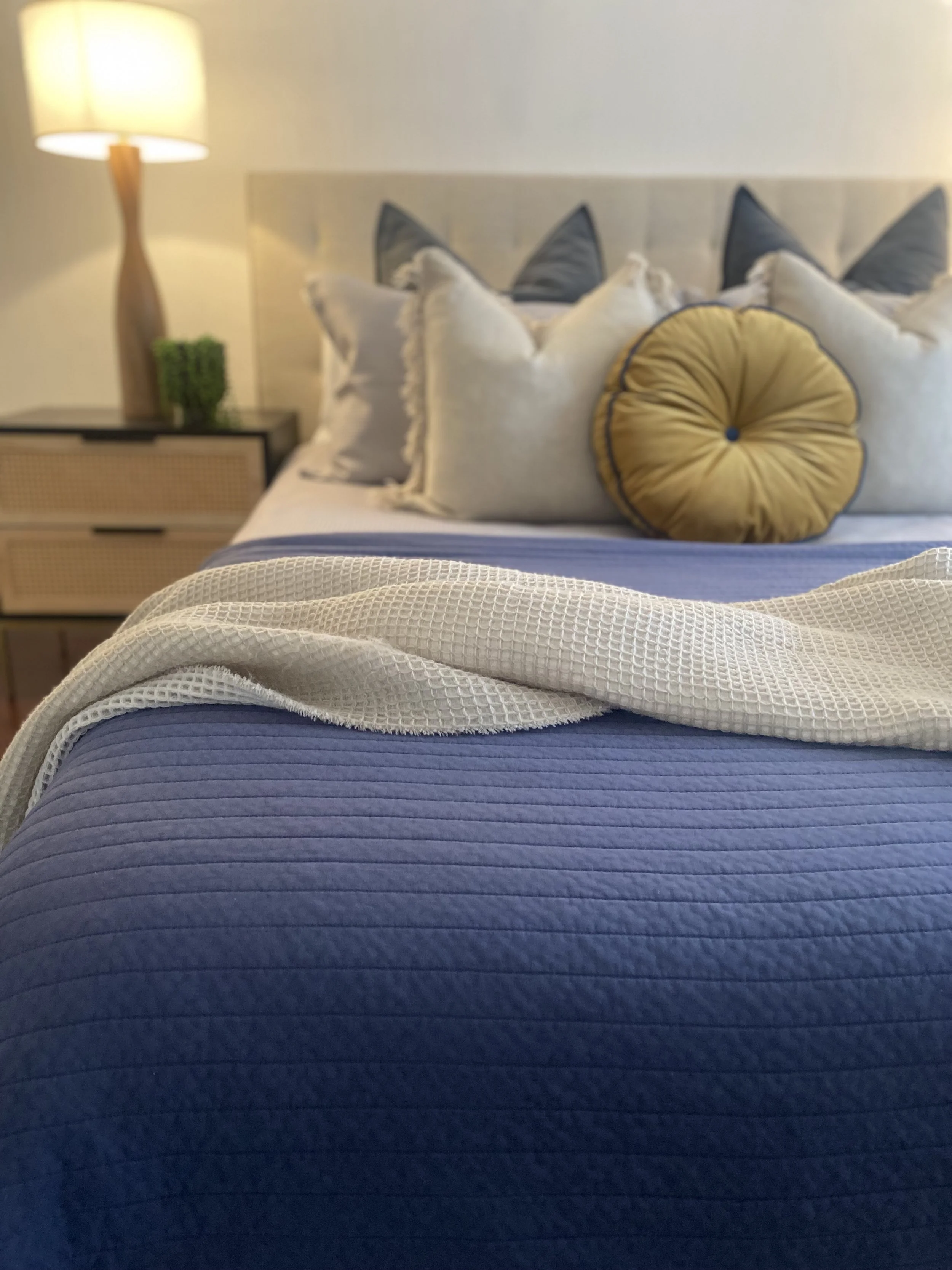 Close-up of a bed with a blue quilt, decorative pillows, and a white throw, in a bedroom with a lamp and nightstand in the background.