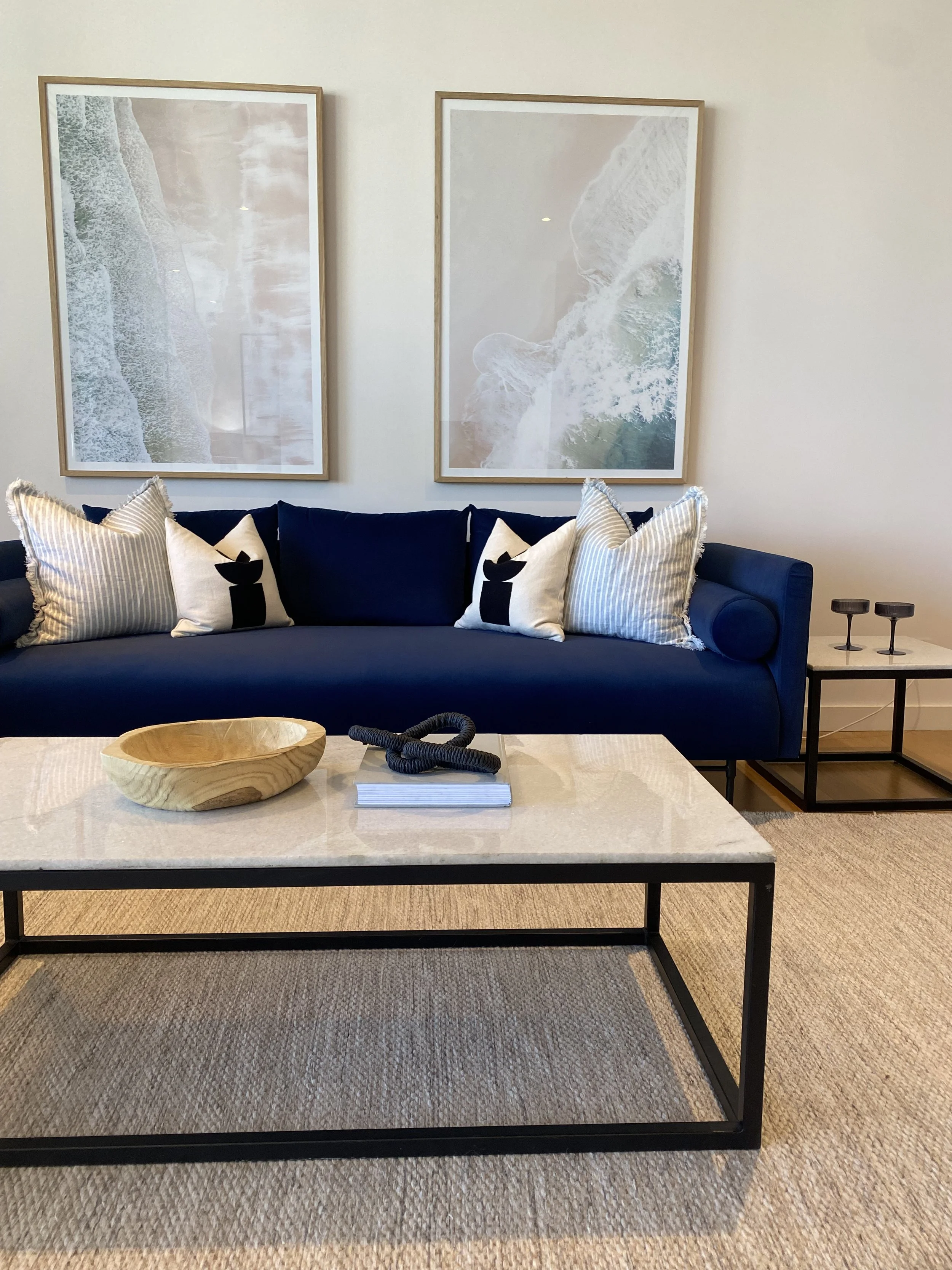 Modern living room with a navy blue sofa decorated with white and blue striped pillows, large aerial ocean artwork on the wall, a cream-colored marble coffee table with a wooden bowl and decorative rope sculpture, and a side table with two black cand