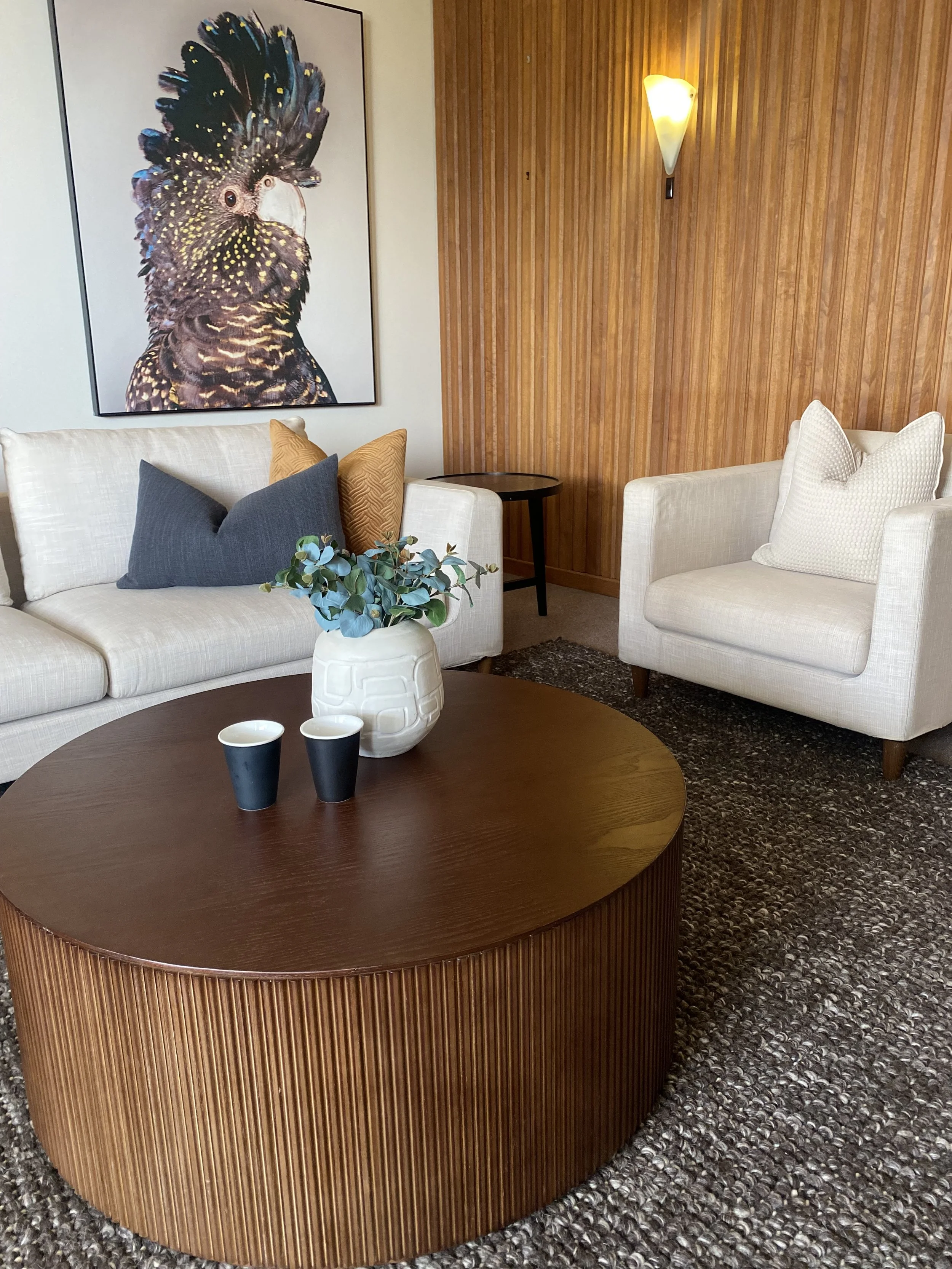 A living room with a wooden-paneled wall, a framed picture of a cockatoo, a cream-colored sofa with cushions, a round wooden coffee table with a white vase and two dark cups, a cream armchair with a textured pillow, a small black side table, and a wa