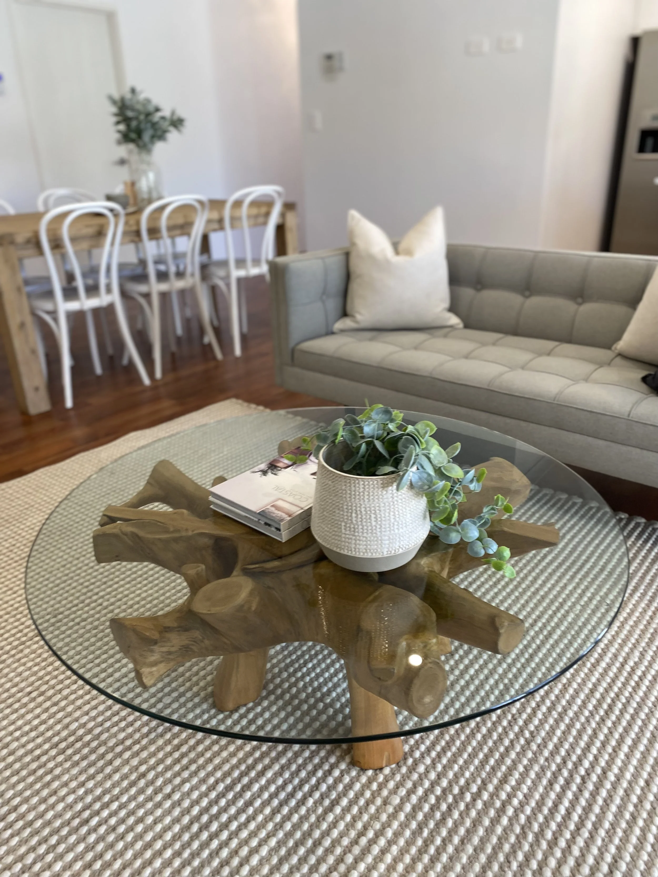 Living room with a wooden coffee table with a glass top, a plant, and magazines, a beige tufted sofa with pillows, and a dining area with a wooden table and white chairs in the background.