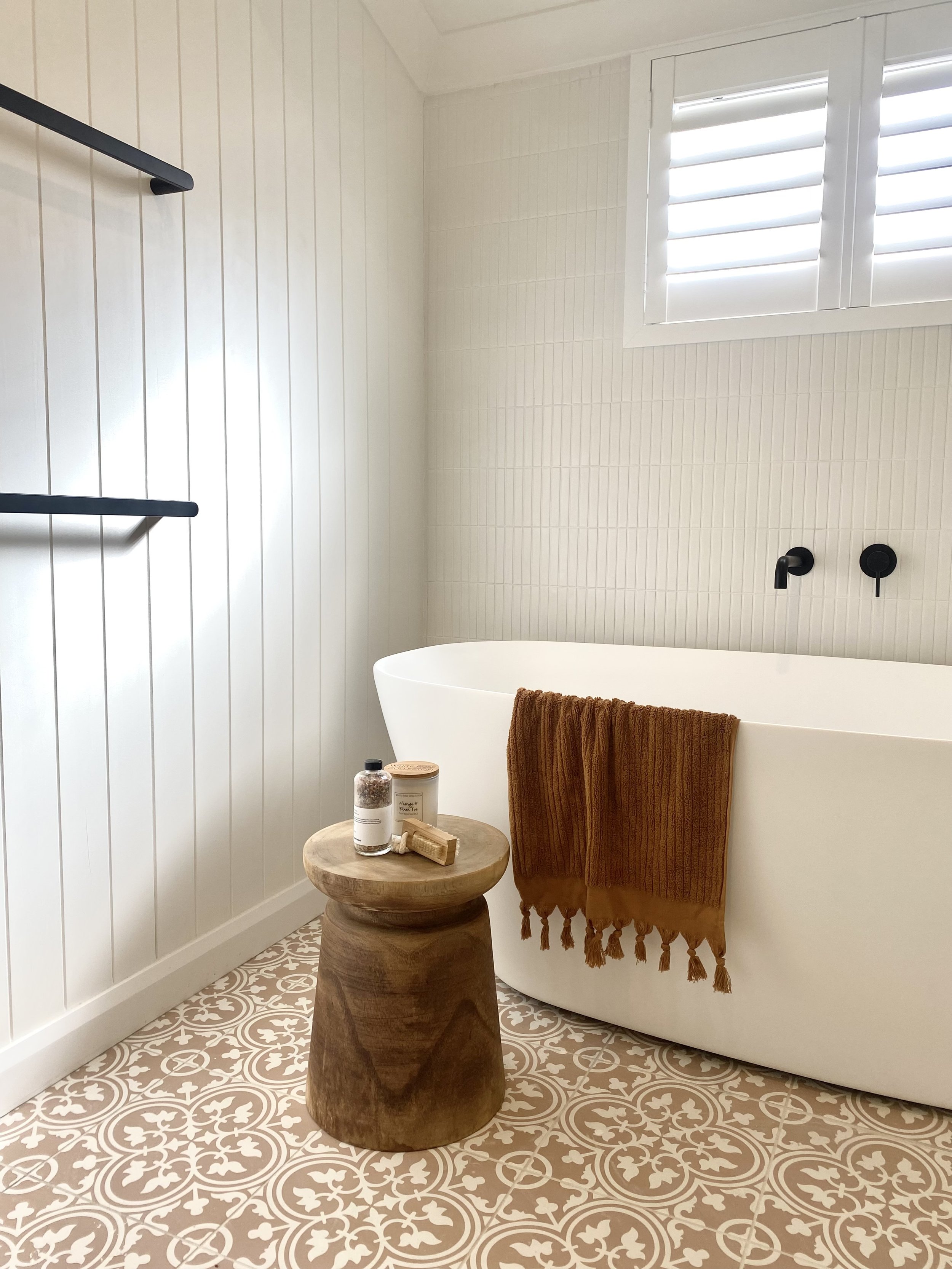 A bathroom with a white freestanding bathtub, a small round wooden side table with bottles and a jar, two rust-colored towels hanging over the bathtub, a window with white shutters, and patterned beige and white floor tiles.