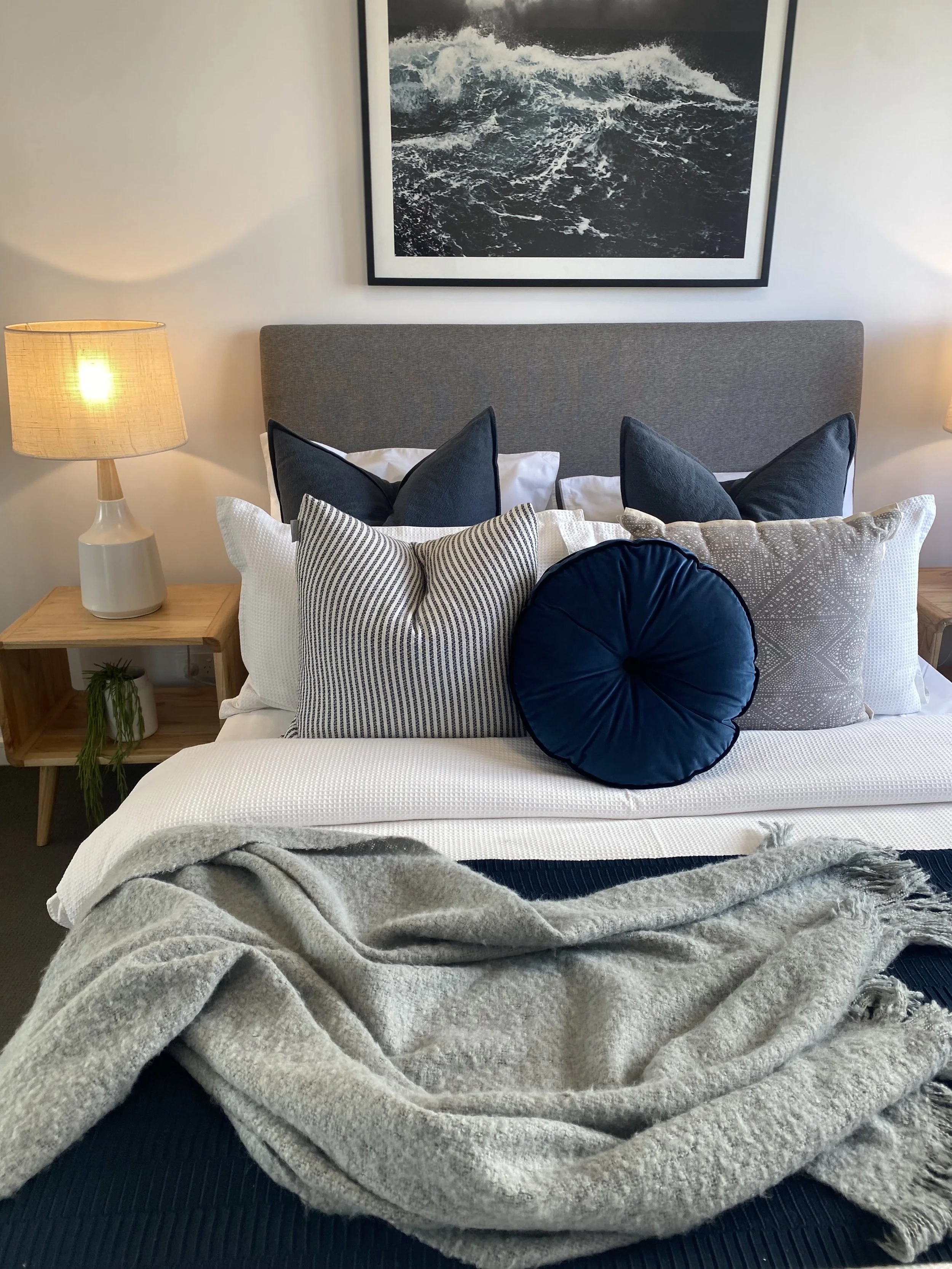 A neatly made bed with multiple pillows, including navy blue, patterned, and round decorative pillows, topped with a cozy beige blanket, in a bedroom with wooden nightstands, lamps, and a framed ocean photograph on the wall.