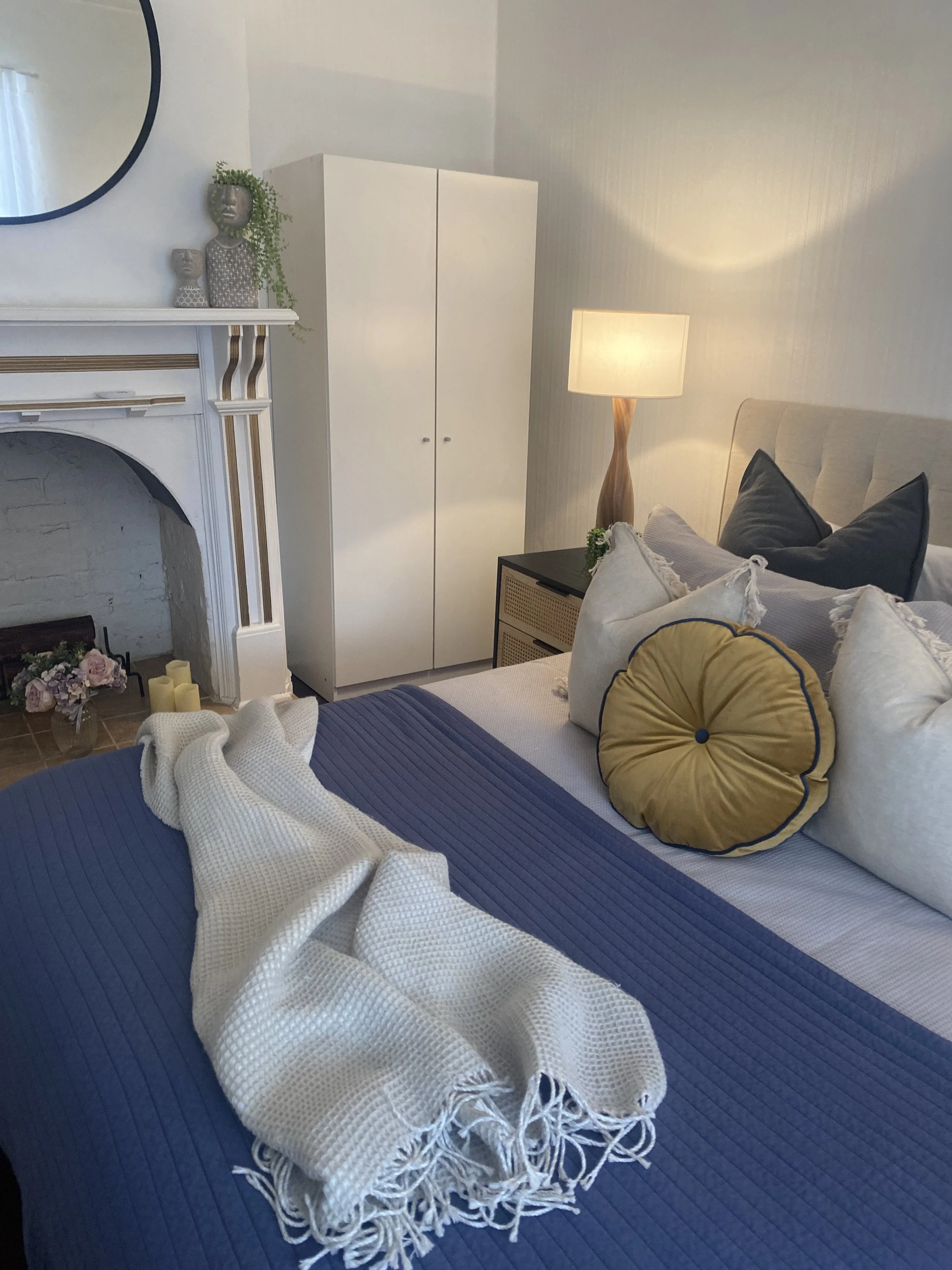 Cozy bedroom with a bed covered in a blue blanket, beige pillows, and a white throw blanket. A bedside lamp with a wooden base, a white wardrobe, and a fireplace with decorative candles and flowers are visible.