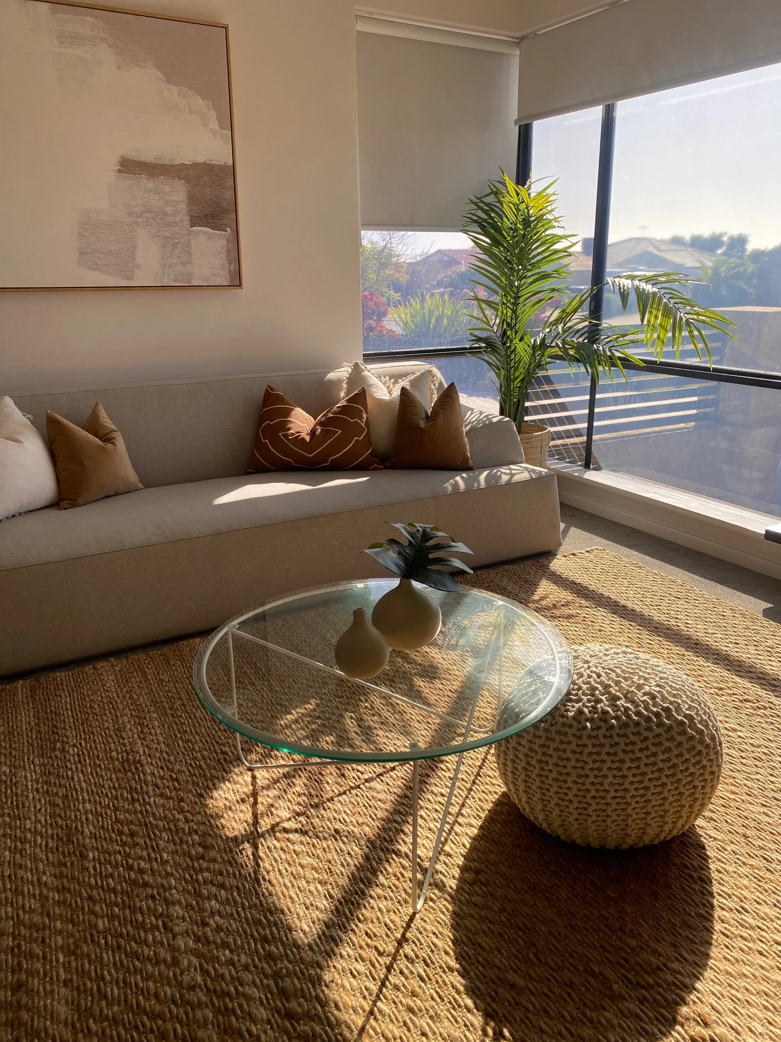 Living room with beige sofa, decorative pillows, round glass coffee table with two vases, knitted pouf, large window with blinds, potted plant, wall art, and textured rug.