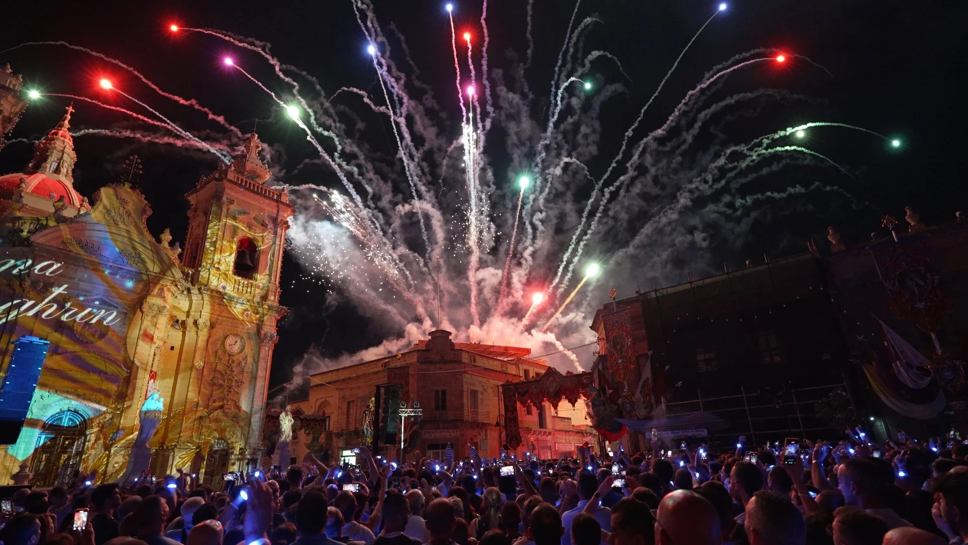 Nighttime celebration with fireworks over historic buildings, crowd of people taking photos and videos.