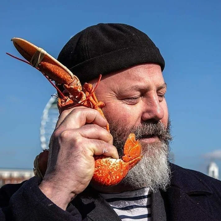 A man with a beard and black beanie holding a lobster to his ear, as if talking on the phone, against a clear blue sky.