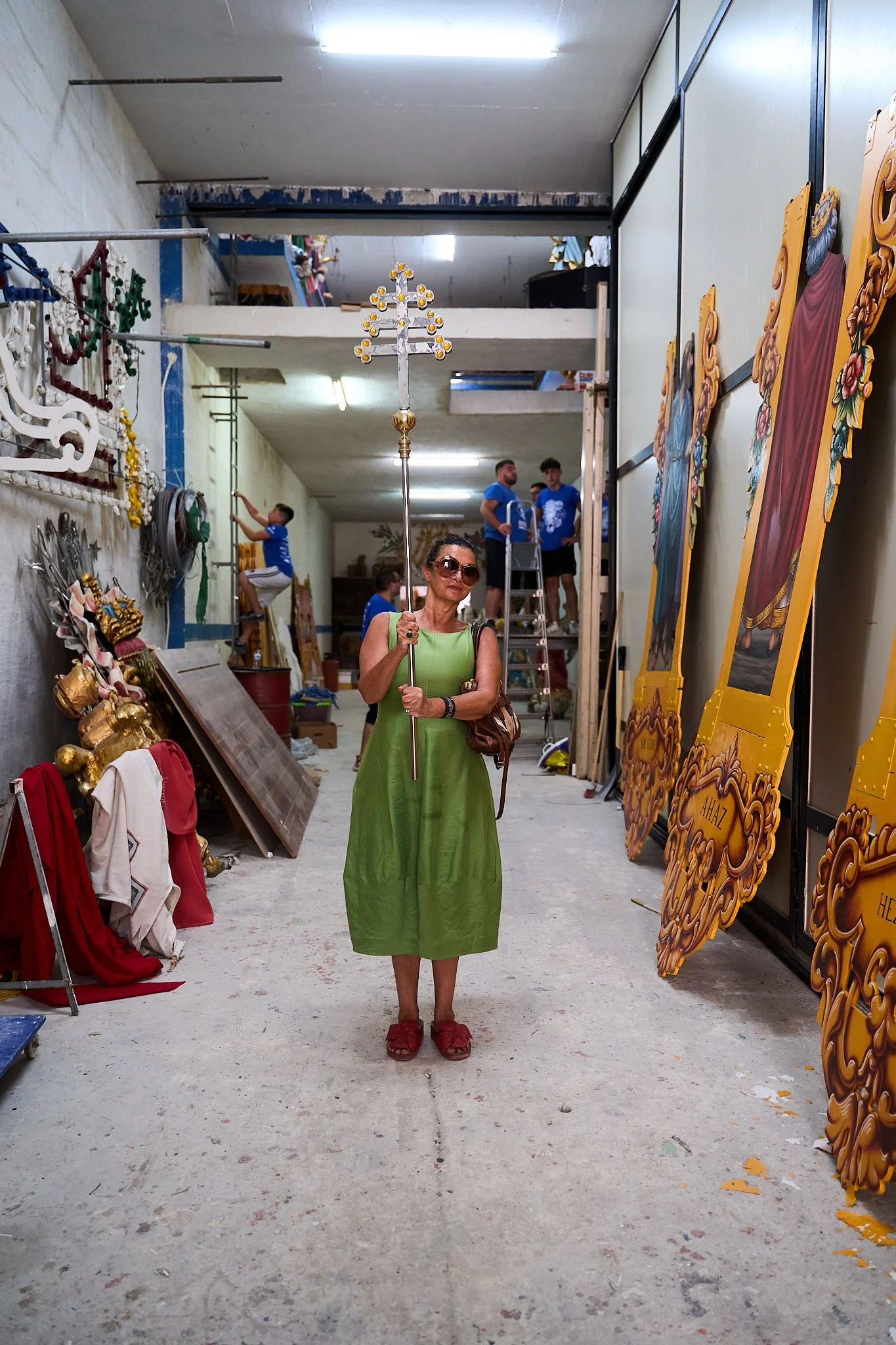 A woman in a green dress and red sandals wearing sunglasses and holding a religious cross inside a workshop or storage area filled with religious statues, landscape panels, and tools, with several people in the background.