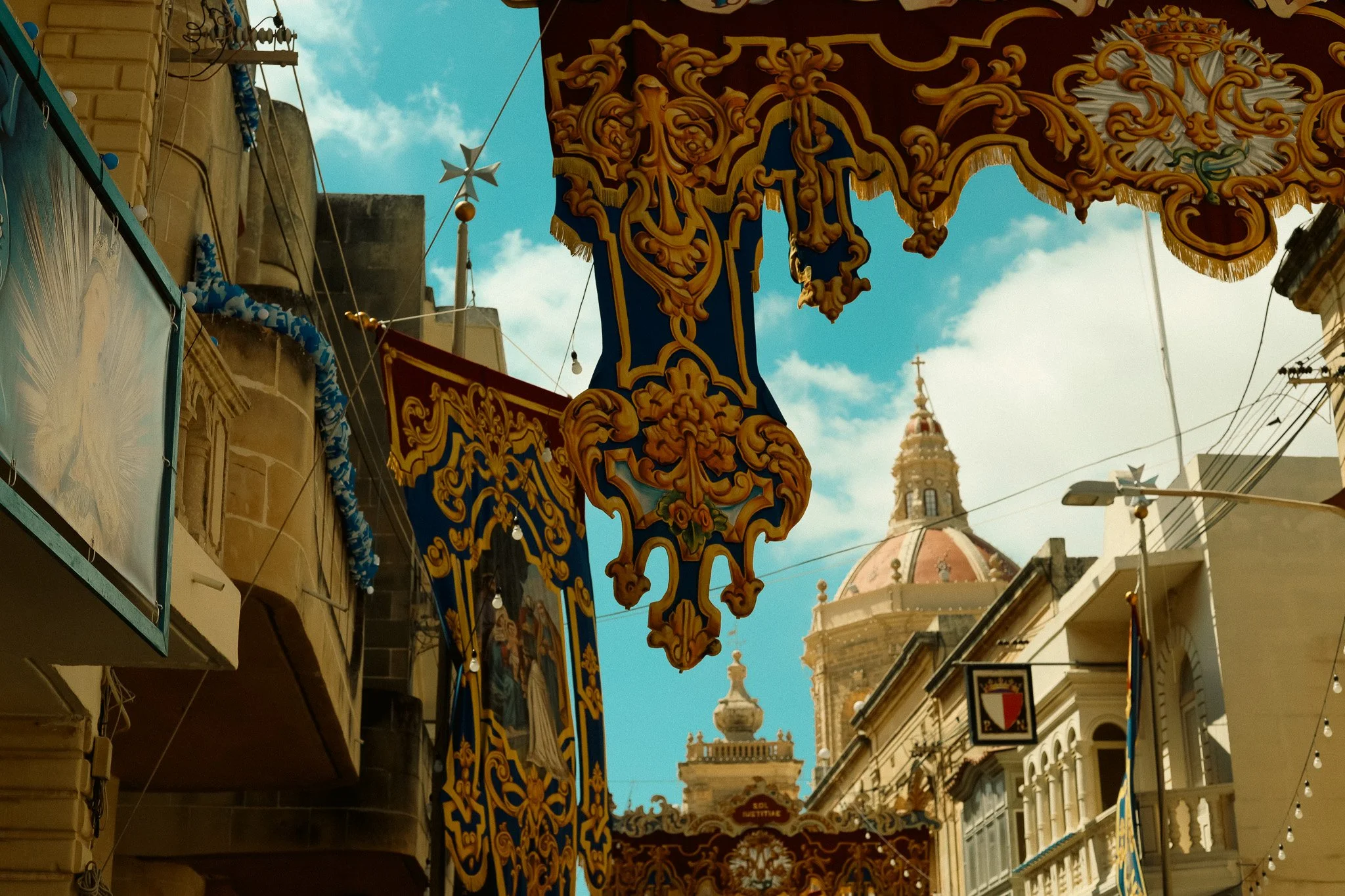 Colorful ornate decorations hang above a narrow street with a church and historic buildings in the background, under a blue sky with clouds.