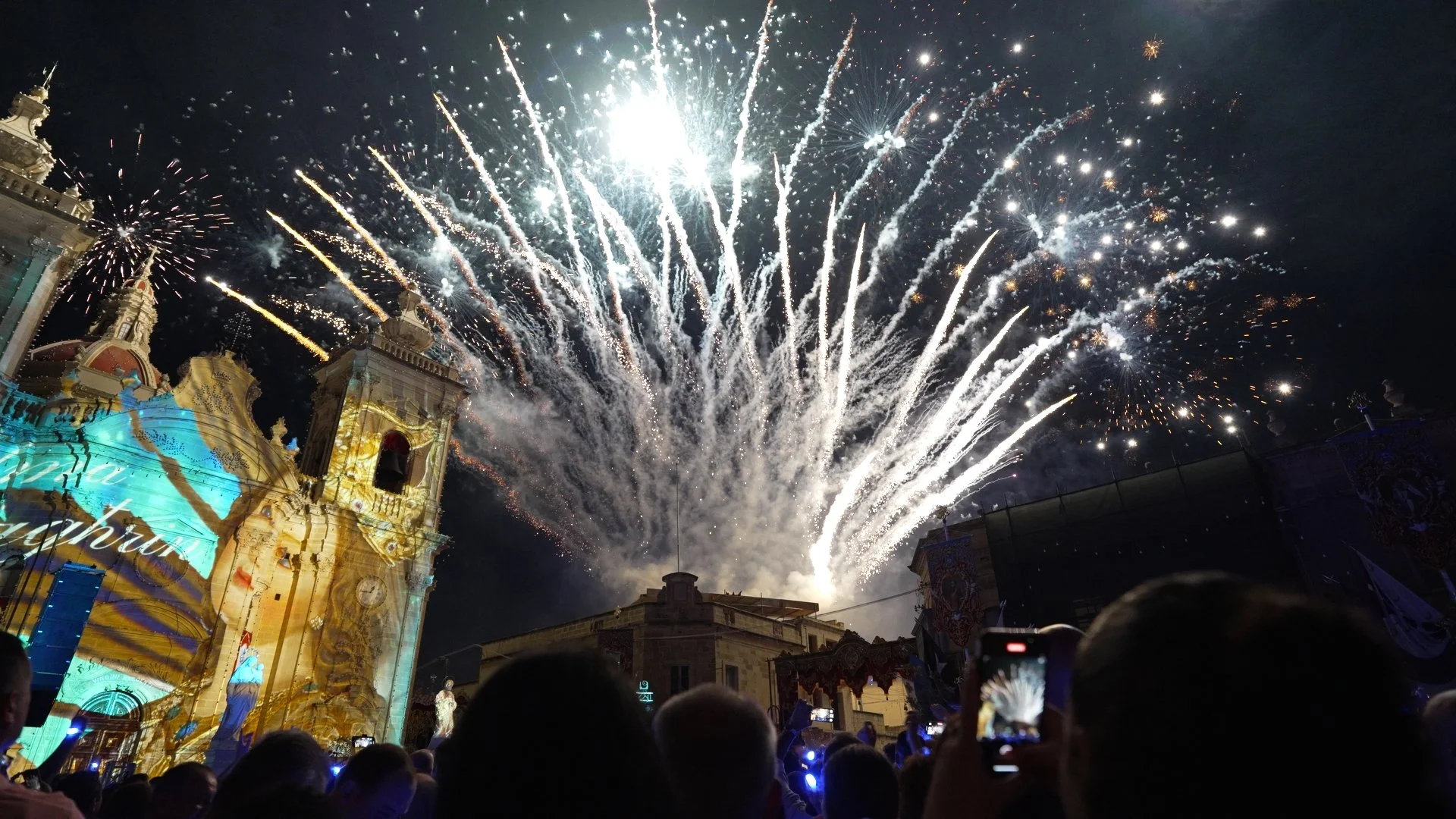 Fireworks explode above a historic church during a celebration at night, with a crowd of people watching and taking photos.