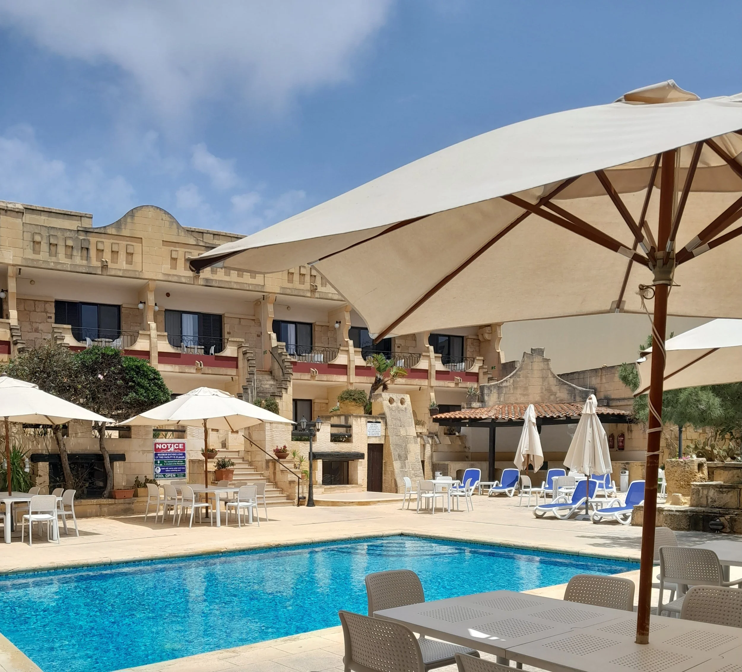 Swimming pool area at a hotel with white umbrellas, lounge chairs, and a multi-story building in the background under a blue sky with some clouds.