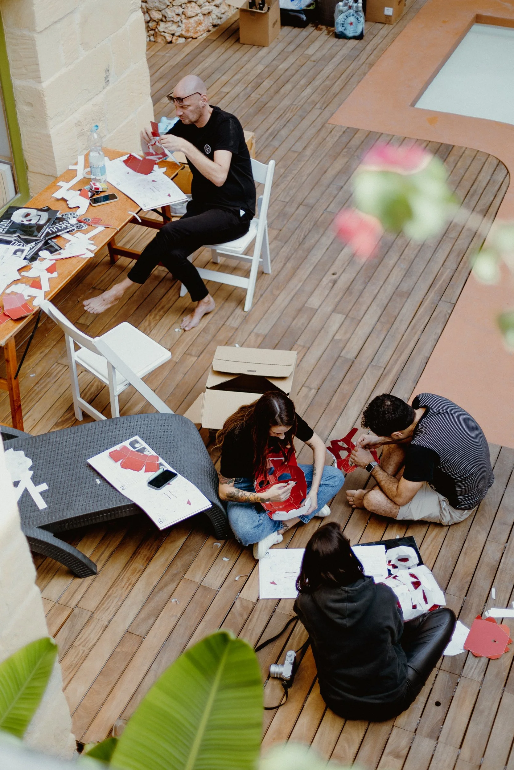People sitting on a wooden patio, working on assembling red and white paper or cardboard crafts, surrounded by instruction papers and boxes - Art Retreat in Malta.