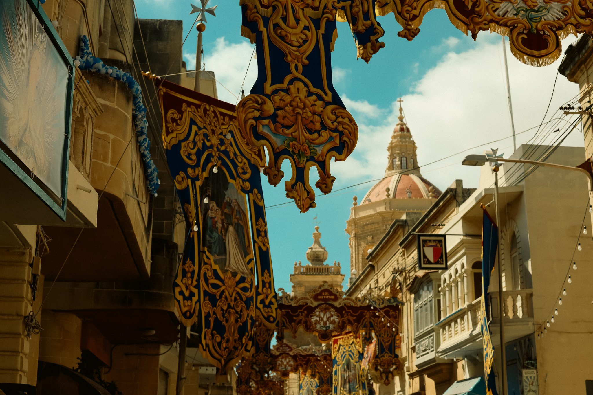 Decorative banners with religious images hanging across a street, with historic buildings and a church dome visible in the background. Art Retreat in Malta.