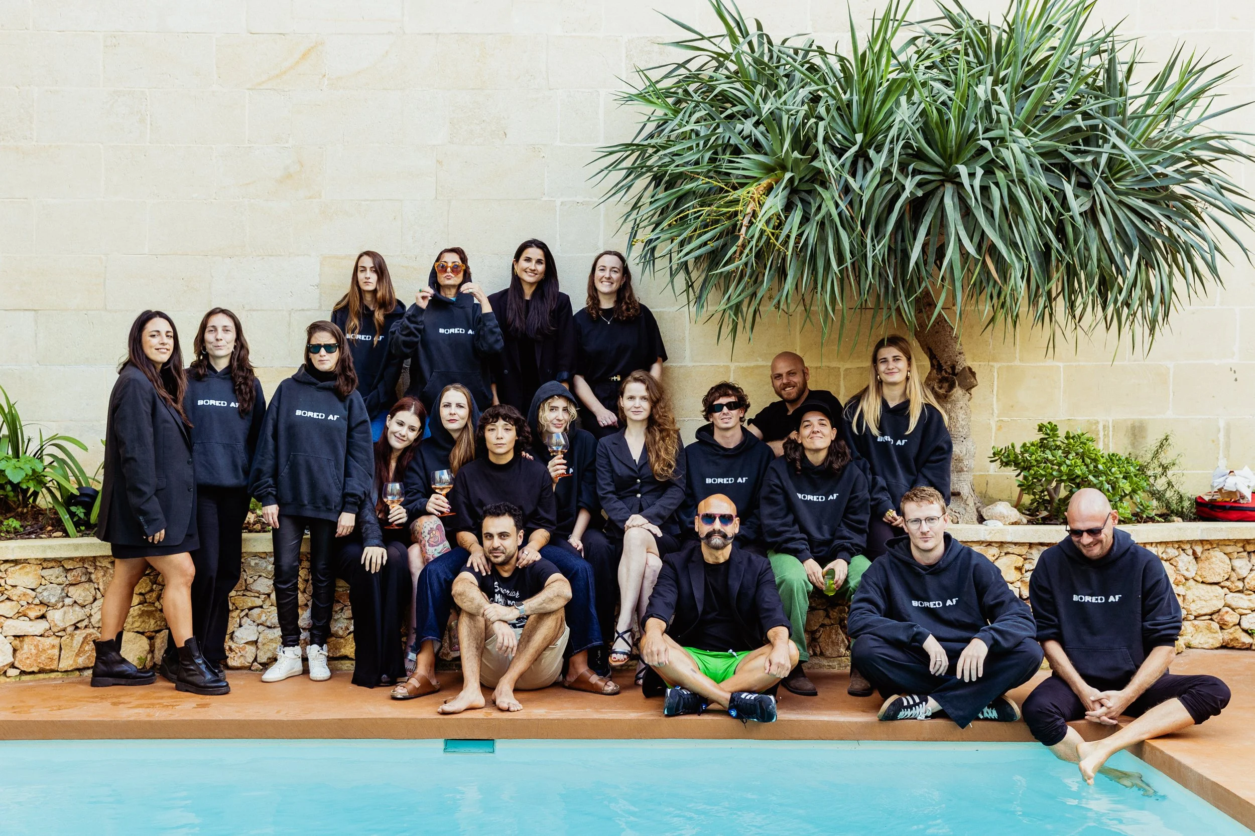 Group of people gathered near a swimming pool under a large leafy tree, some sitting and some standing, wearing casual and dark-colored clothing, with a few holding wine glasses. Art Retreat in Malta.