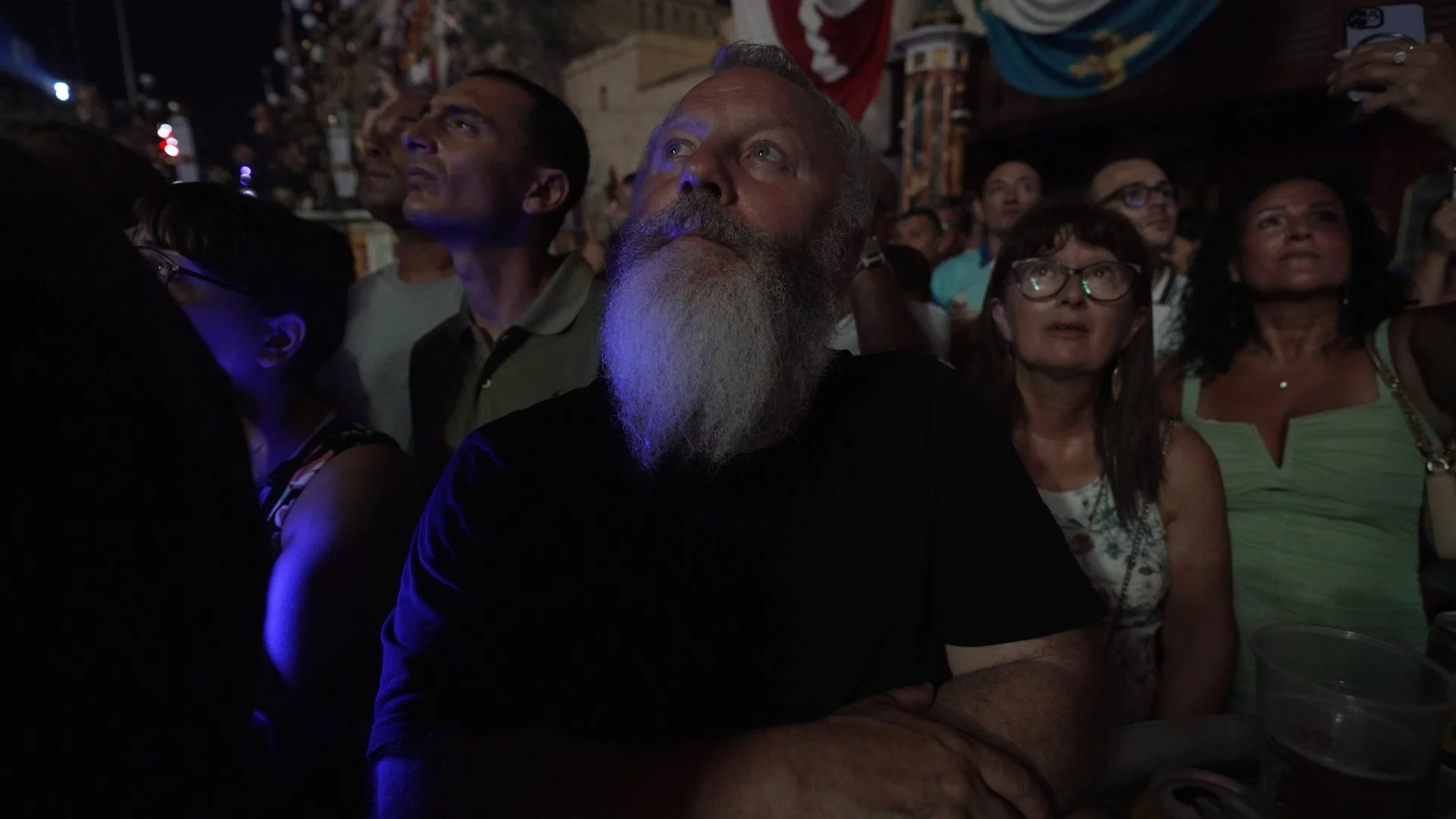 A crowd of people watching a performance or event at night, illuminated by blue and ambient lighting.