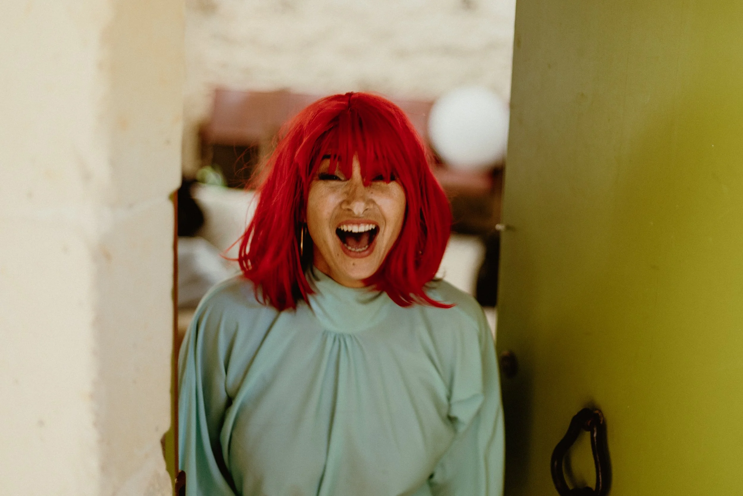 Woman with bright red hair wearing a light blue top, smiling and laughing, standing between a yellow wall and a white wall, with a blurred background.