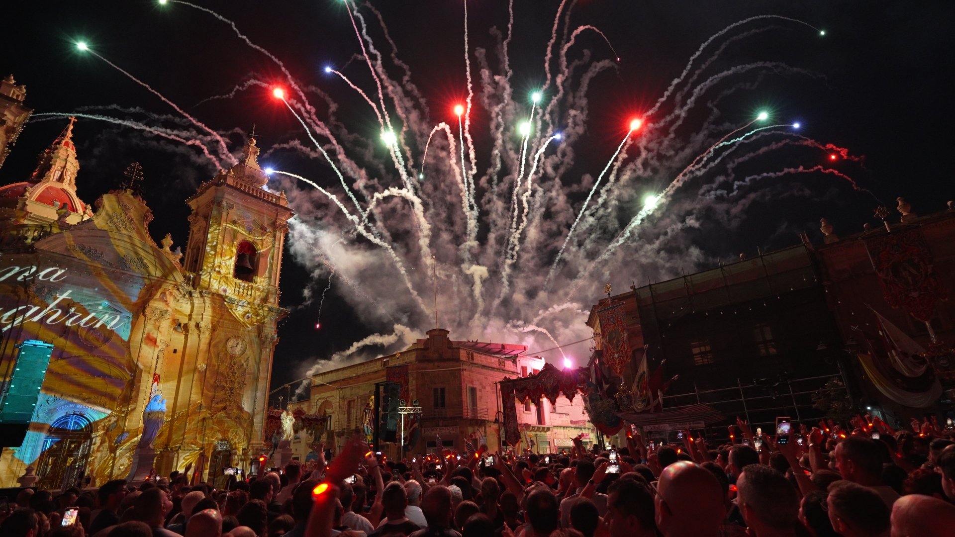 A large crowd watching a fireworks display at night in a historic town square with ornate buildings illuminated, fireworks exploding in the sky.