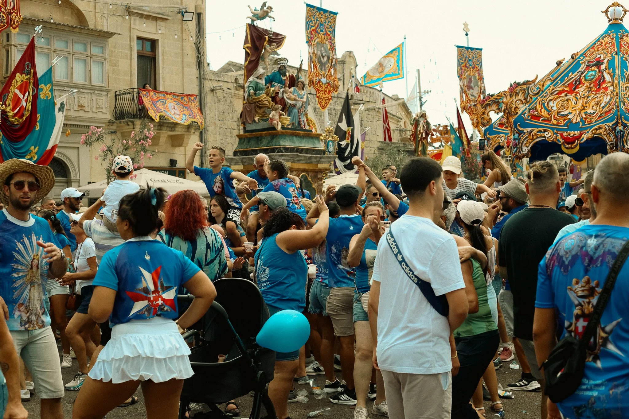 Art Retreat in Malta. Crowd of people celebrating on a street during a festival, with decorated banners and a floating platform with statues in the background.