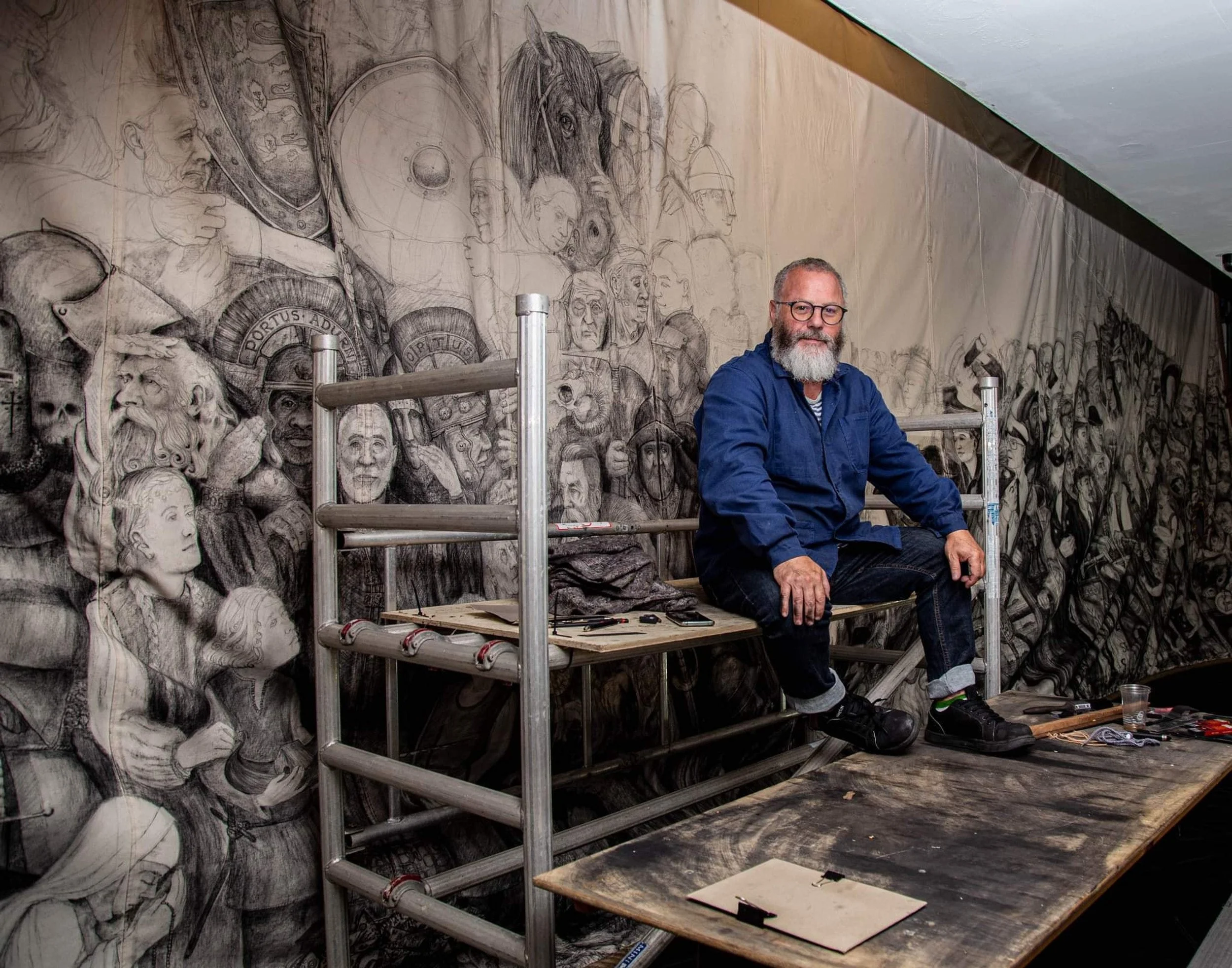 An artist with a beard and glasses, wearing a blue jacket, sitting on scaffolding in front of a large detailed black and white mural featuring faces, animals, and historical figures. Various art tools are on a wooden table nearby.