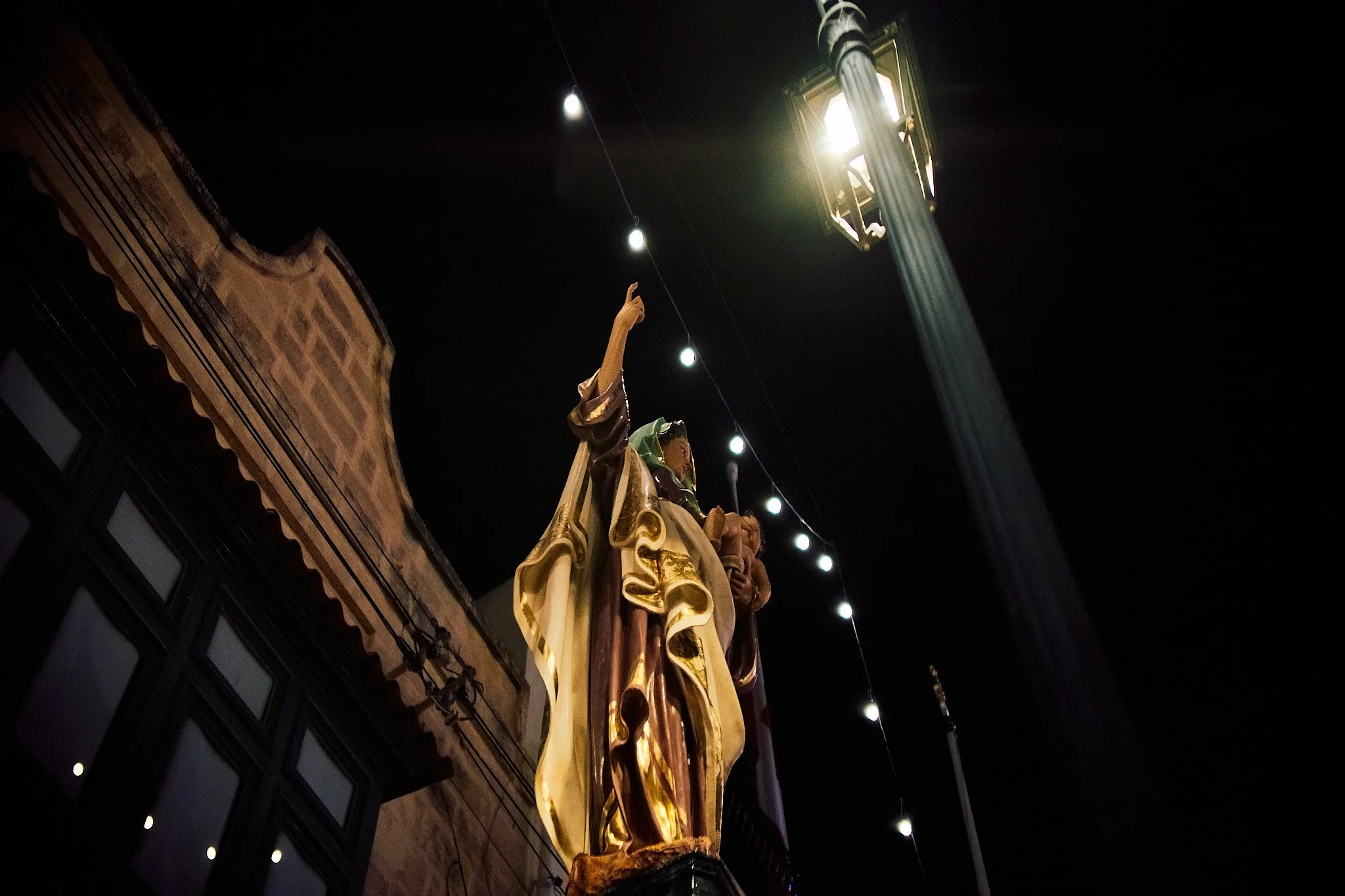 Nighttime view of a religious statue illuminated by street lights, with string lights and an old building in the background.