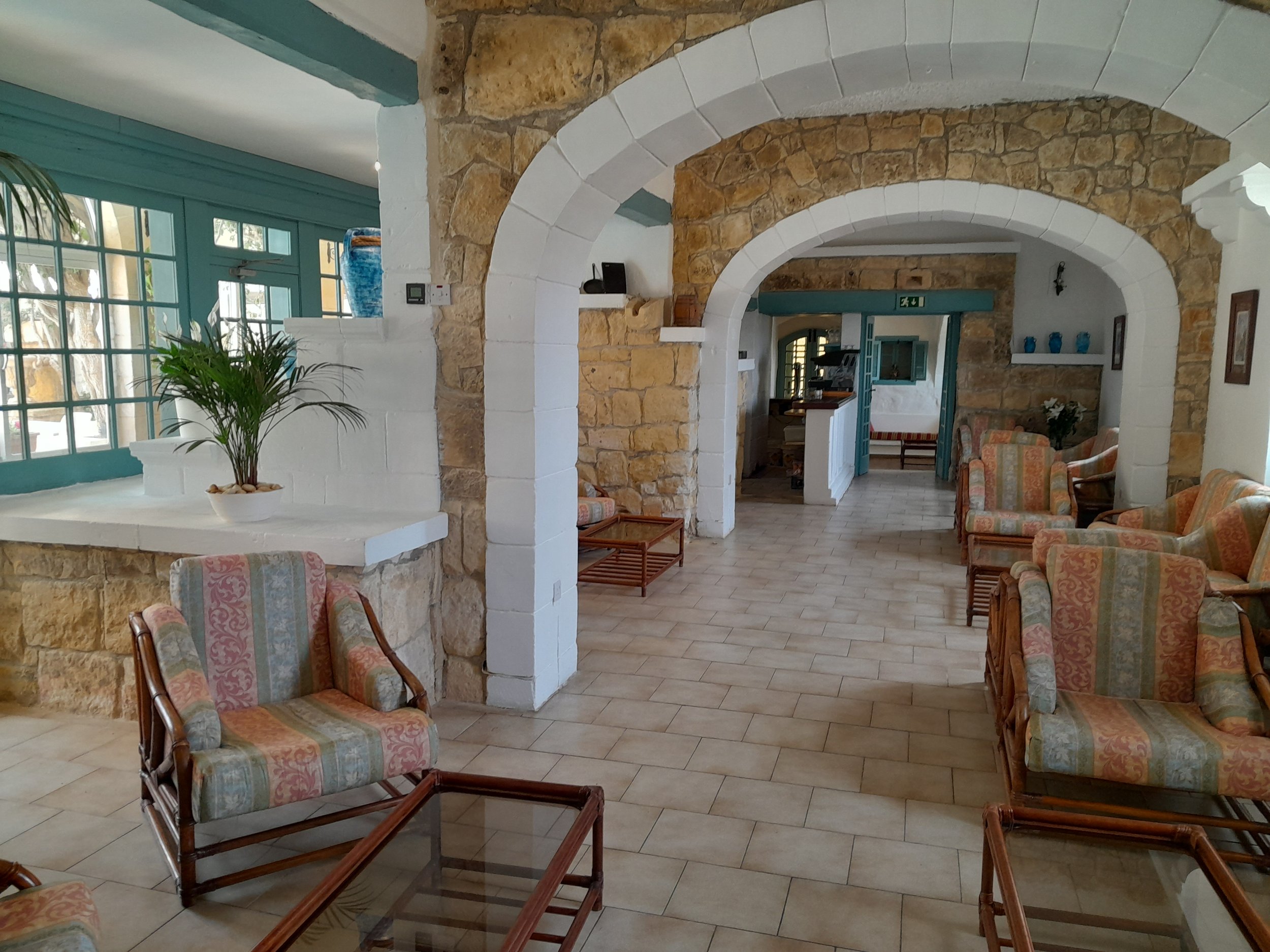 A cozy lobby with archways, stone accents, and pastel armchairs with floral patterns, large windows with blue-green frames, and a potted plant.