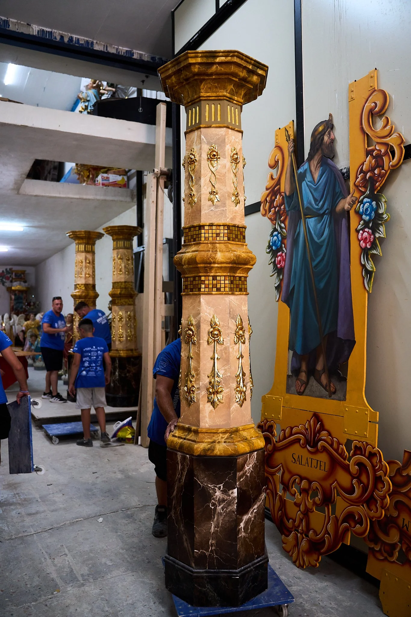 A decorative marble column with ornate gold accents in a workshop surrounded by other similar columns and religious iconography.