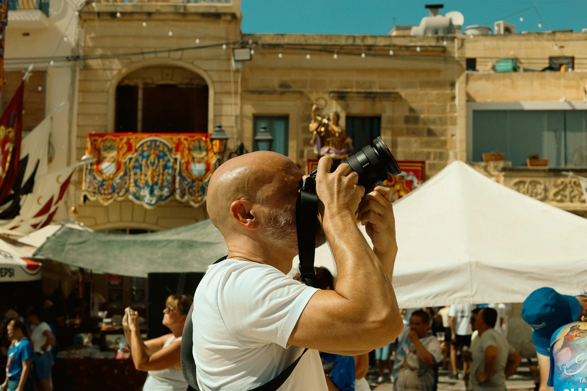 Bald man taking a photograph with a camera in a crowded outdoor setting with tents and decorative buildings in the background.