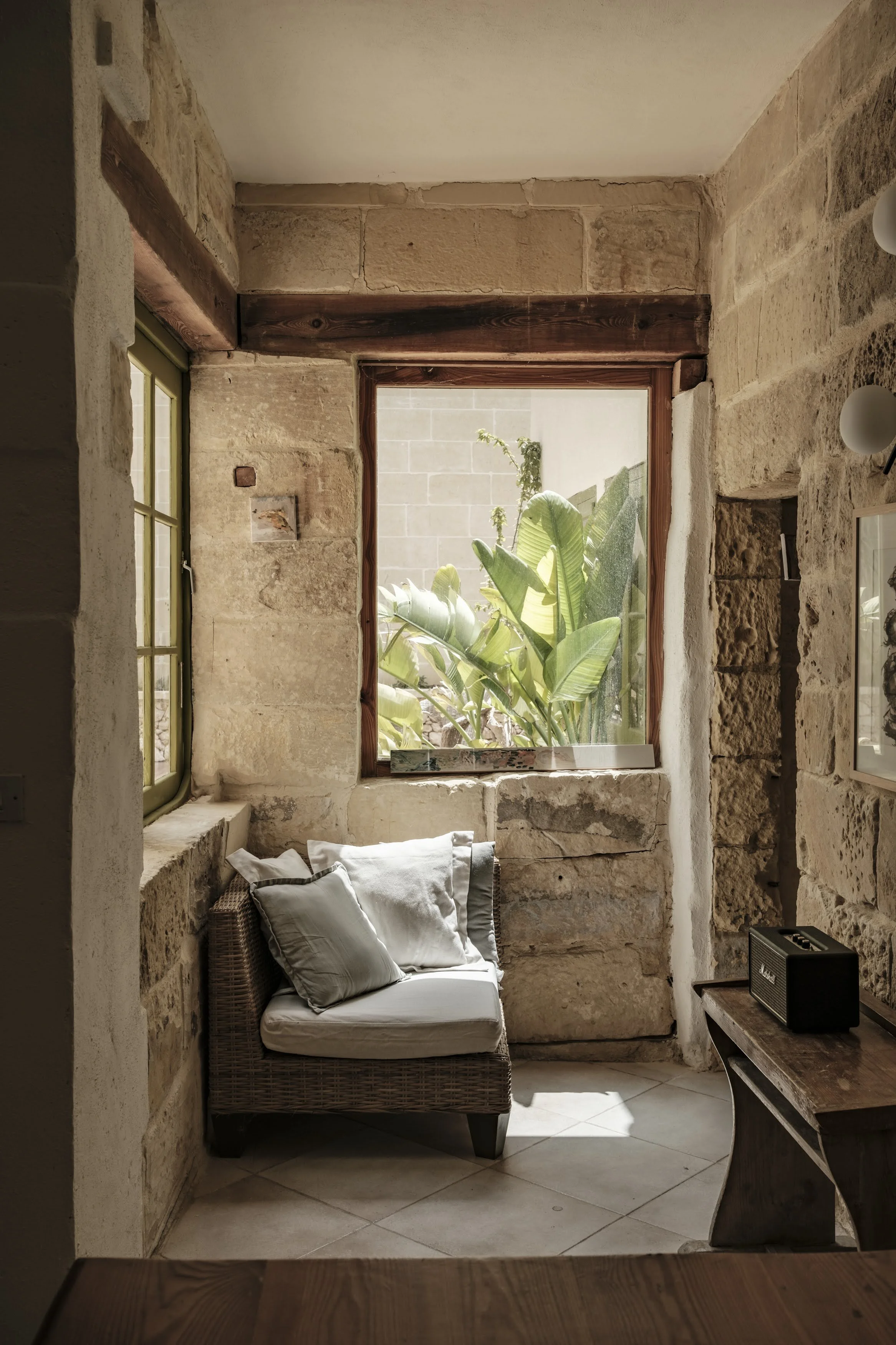 Sunlit corner of a rustic room with stone walls, a wicker chair with cushions, a window with lush green plants outside, and a small wooden table with a radio.
