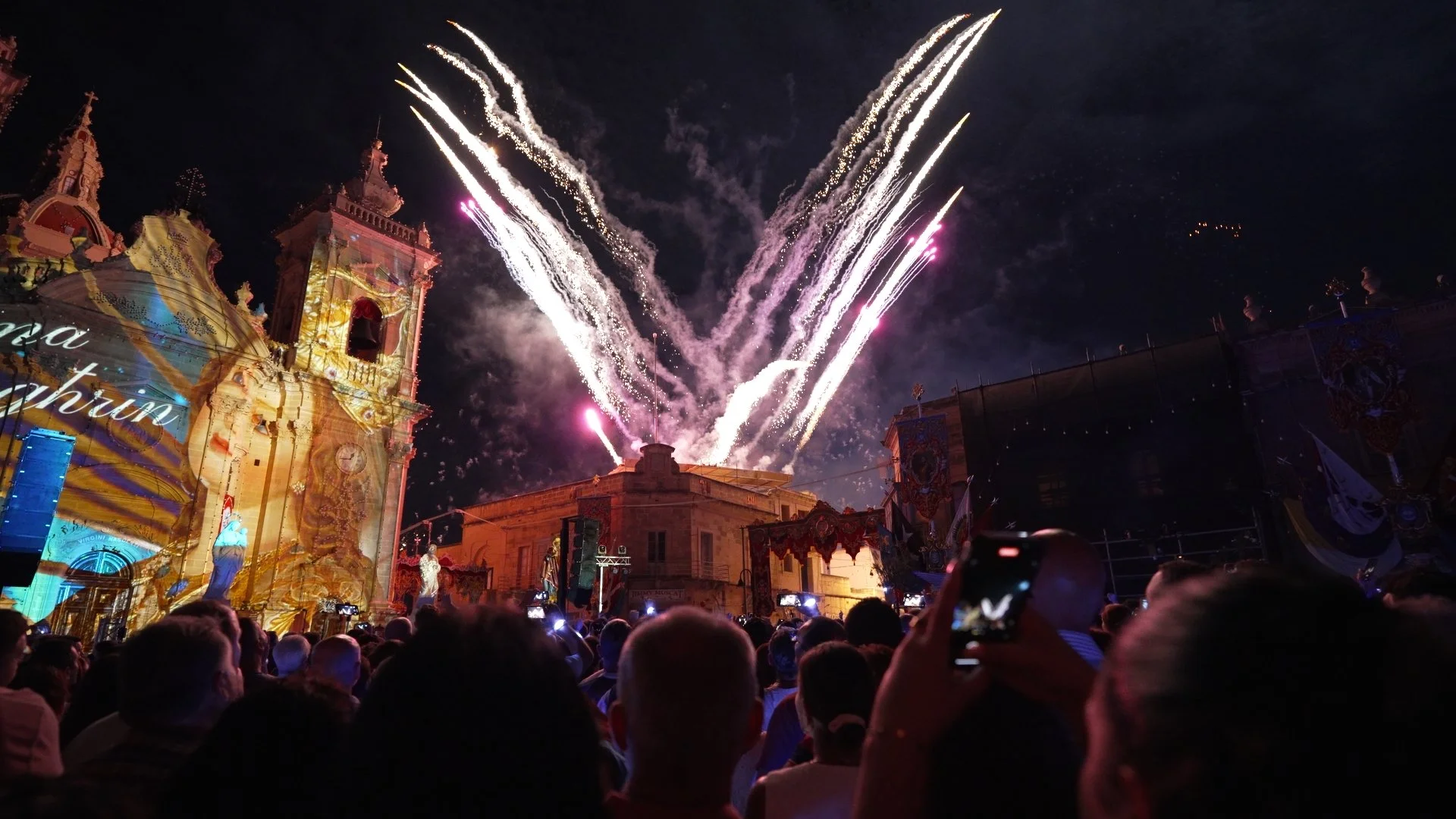 Crowd watching fireworks display over historic buildings at night.